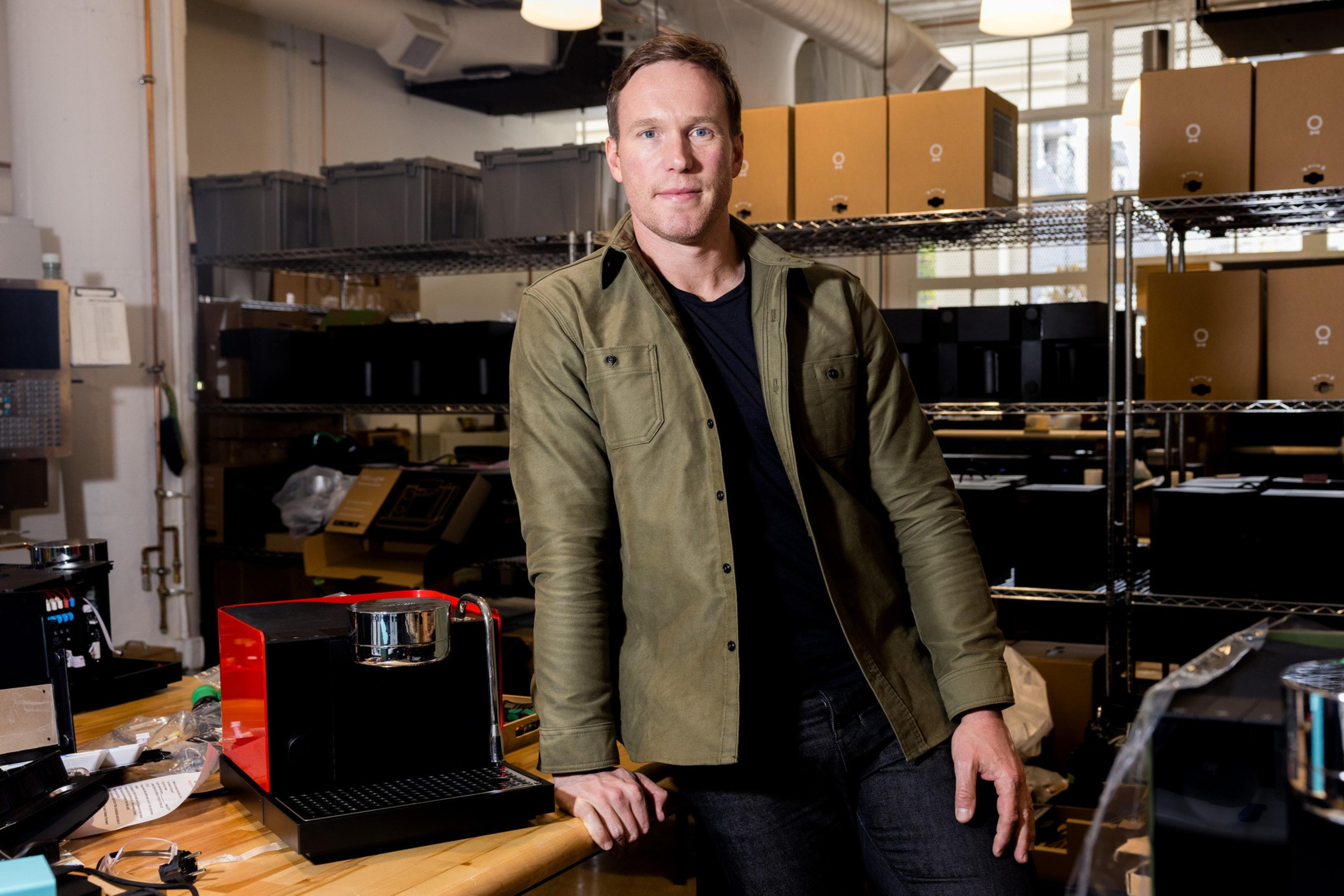 A person in a green jacket is leaning on a workbench beside a red coffee machine. The background has shelves filled with boxes and equipment.