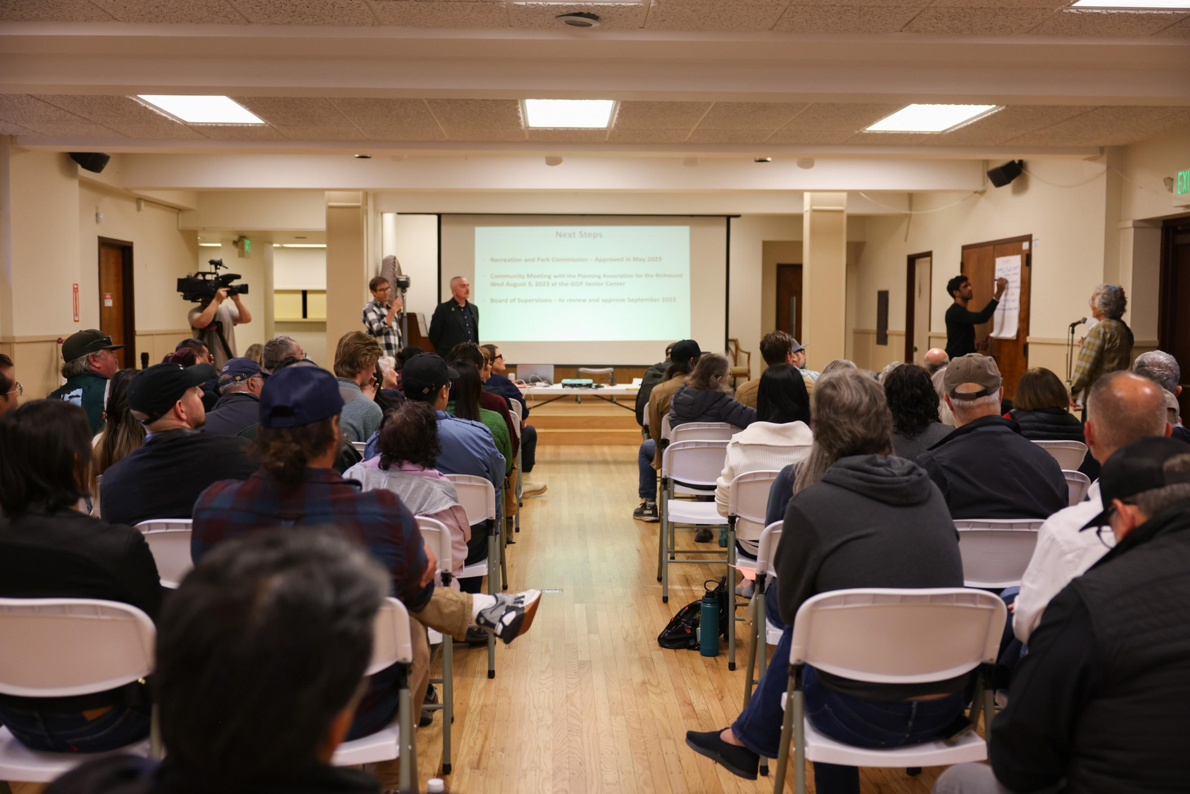 People seated in rows of white chairs face a presentation screen at the front of a room, while two people stand near the screen and one writes on a large pad.