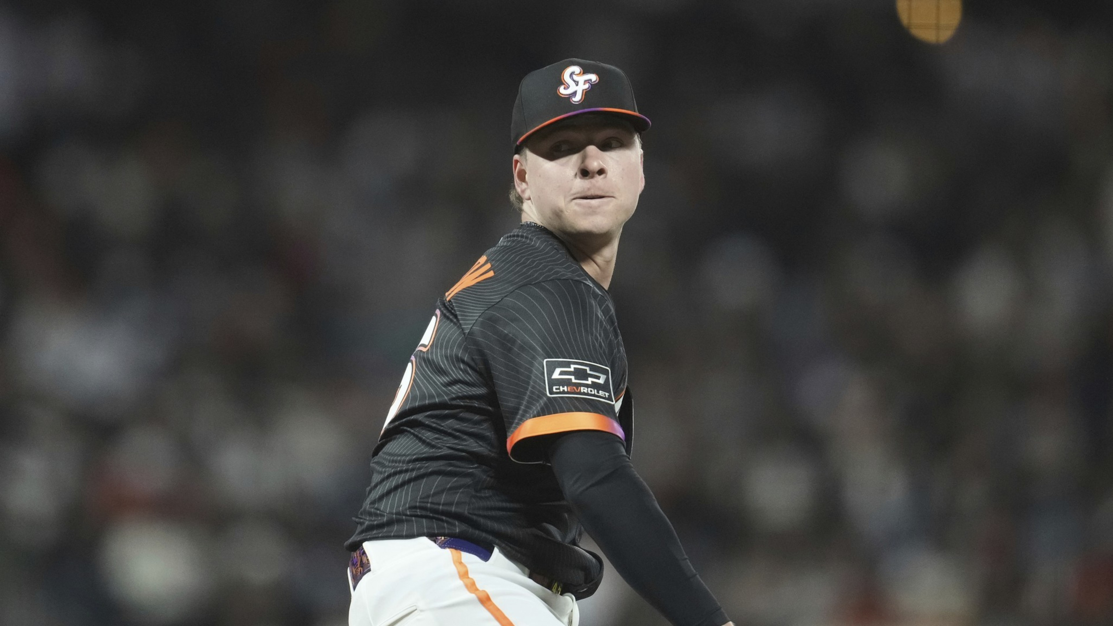 A baseball player is pitching during a game, wearing a black uniform with orange accents and an &quot;SF&quot; cap. The background is blurred with a crowd visible.