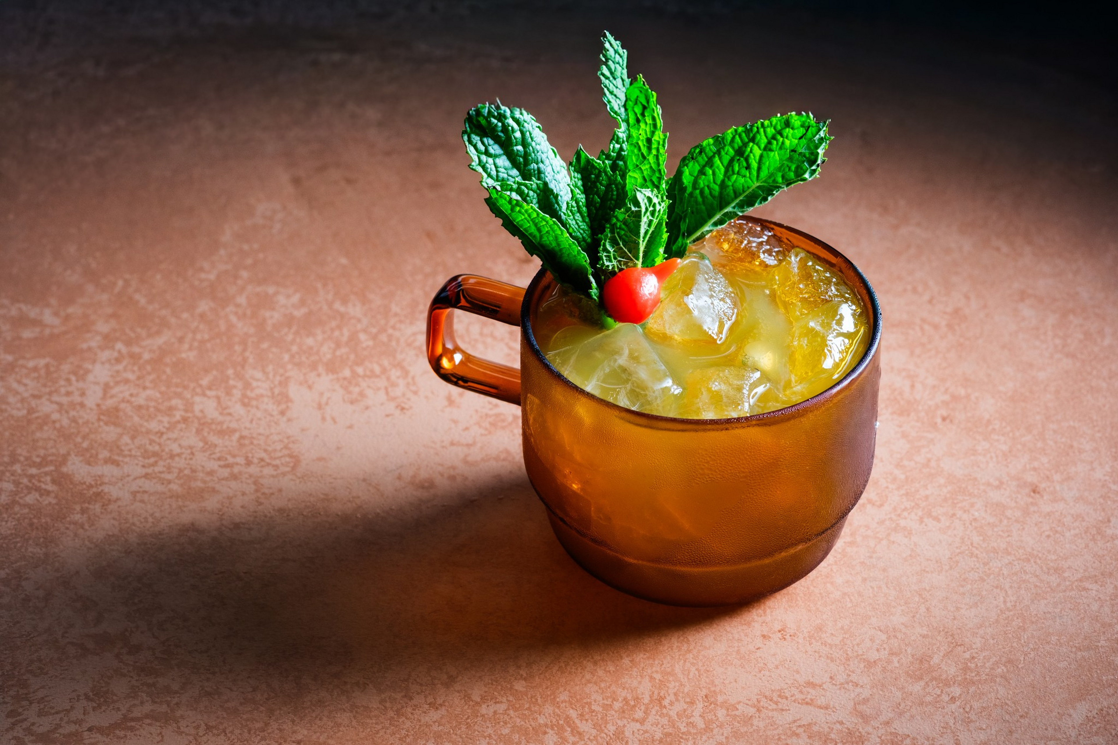 An amber glass mug filled with an iced yellow drink garnished with fresh green mint leaves and a small red cherry on top.