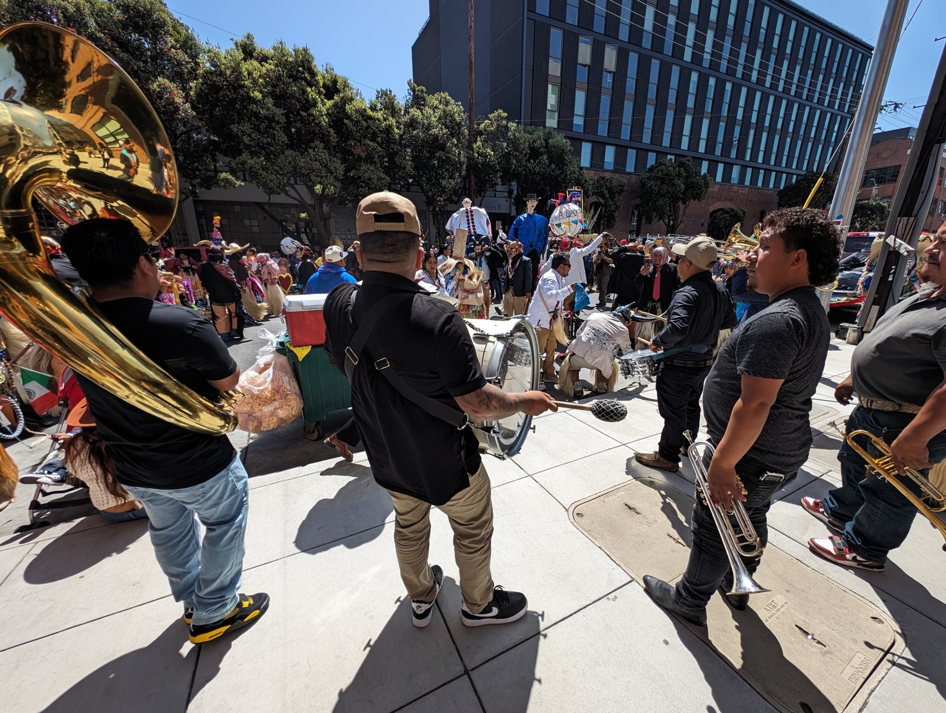 Musicians beat drums and hold horns on a sunlit sidewalk during a parade.