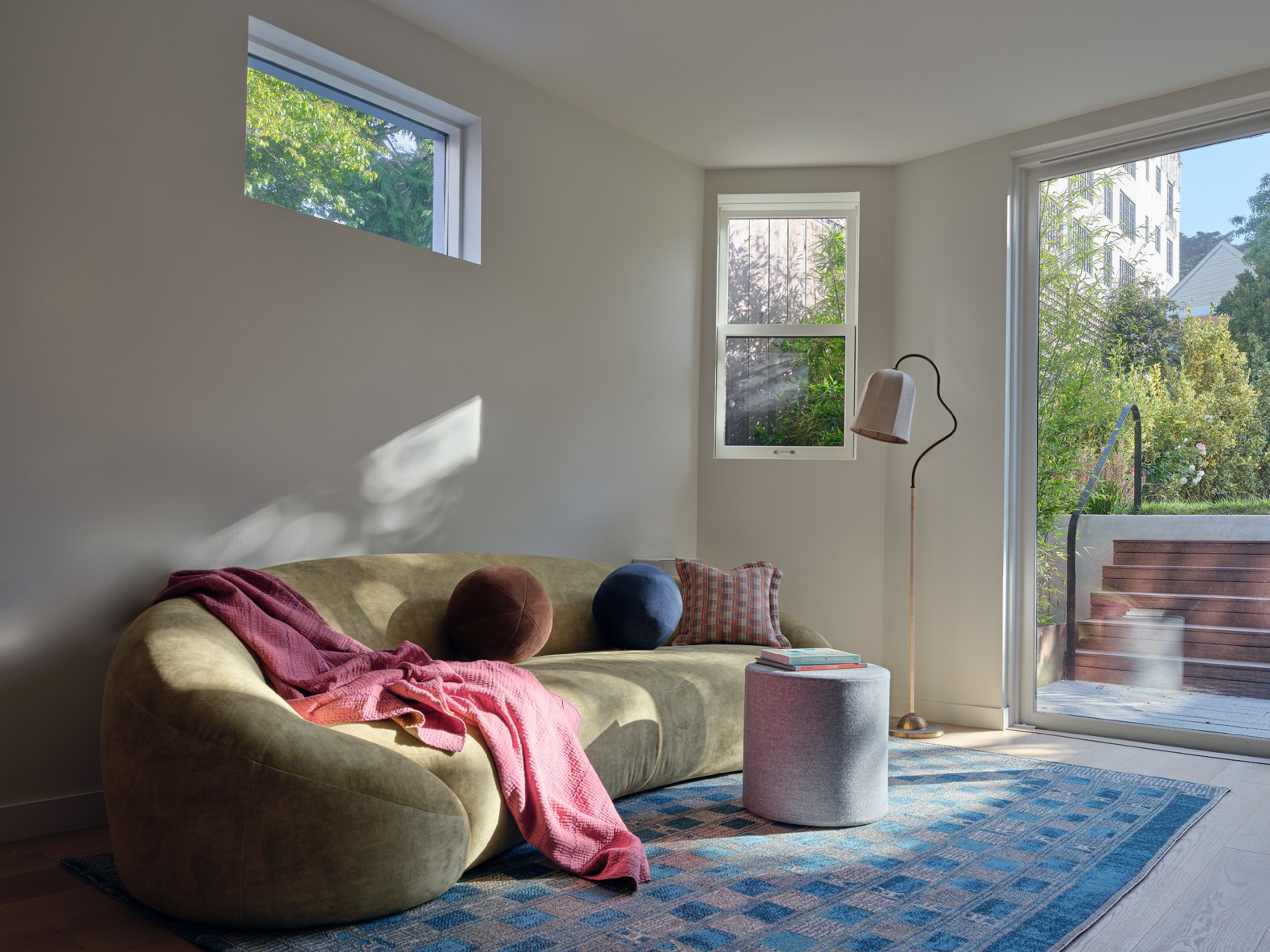A curved olive-green sofa with pillows and a pink throw sits on a blue patterned rug next to a floor lamp and a round ottoman by large windows.