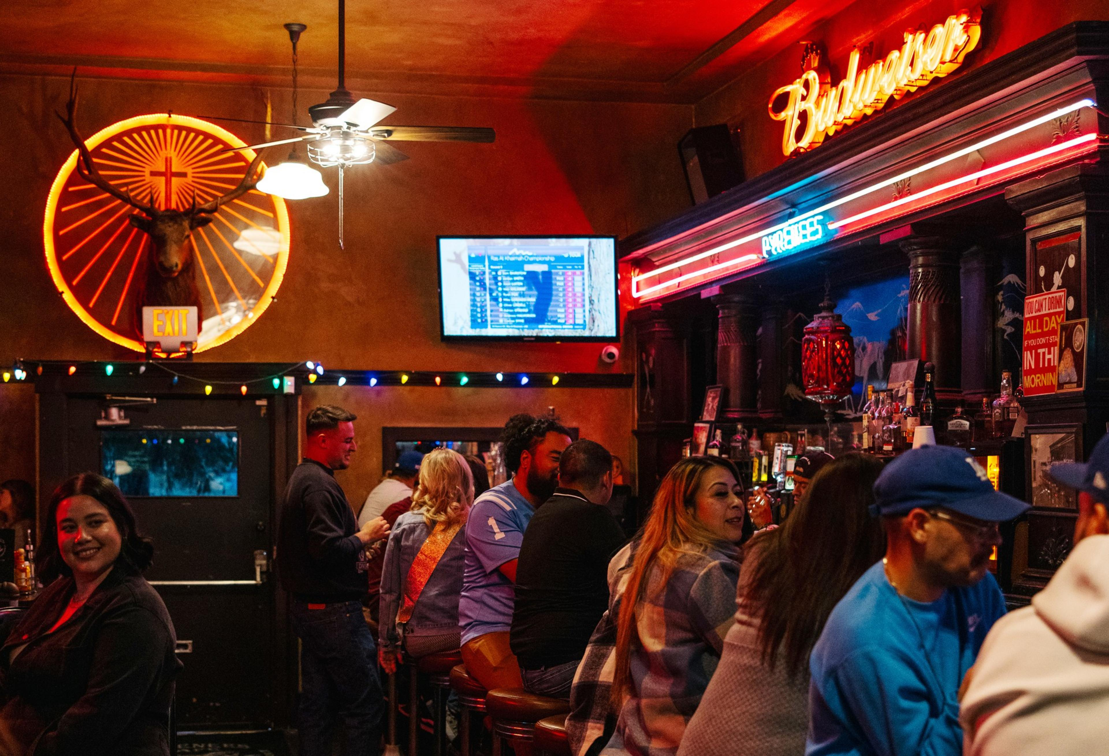 The image shows a lively bar scene with people socializing. A neon Budweiser sign is above the bar, and a glowing deer head decorates the wall.