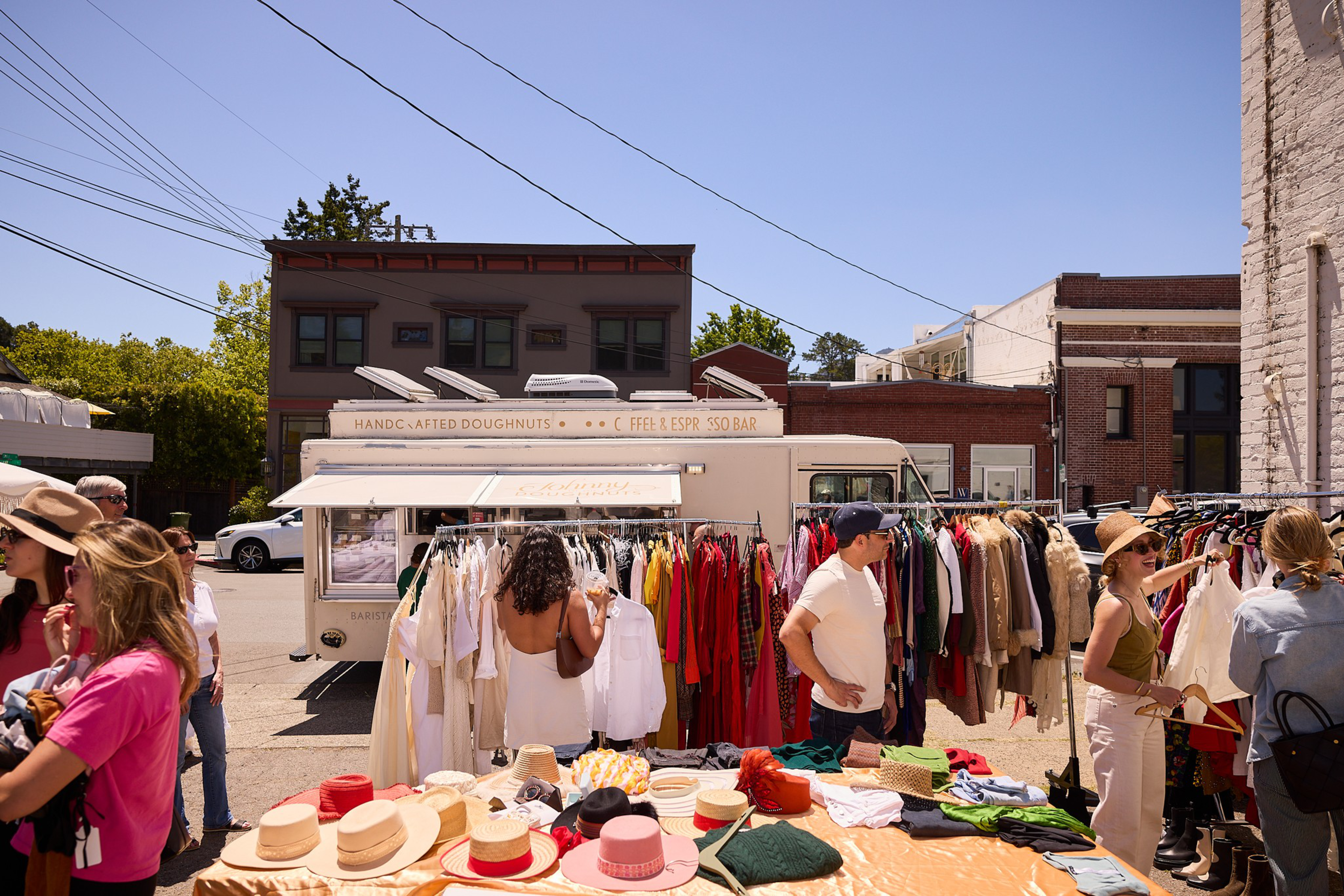 People browse clothing racks and hats at an outdoor market set up in front of a white food truck labeled “Handcrafted Doughnuts.”
