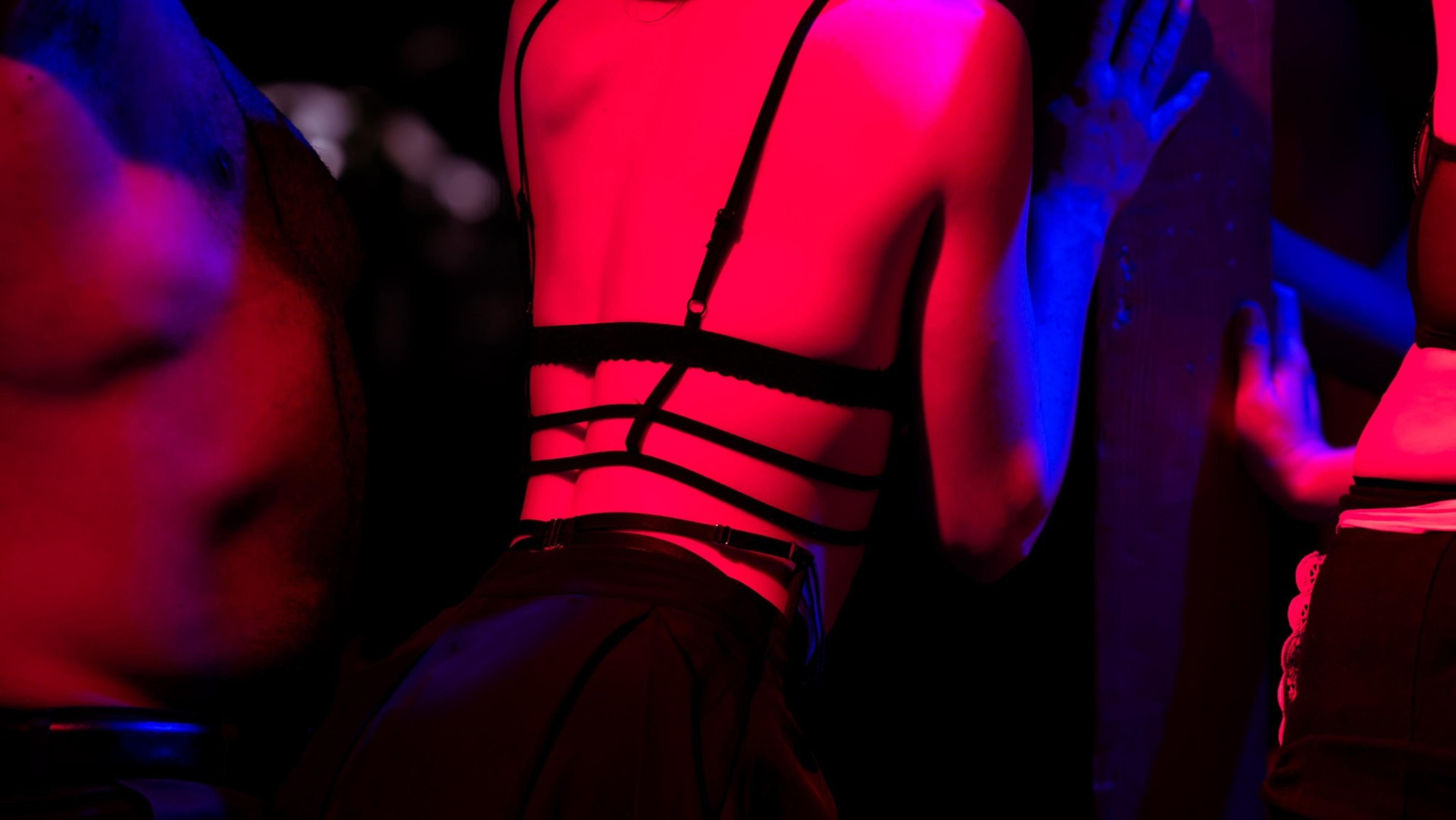 A person wearing a strappy black top presses their hands against a wall, bathed in red and blue lighting, with parts of other figures visible nearby.