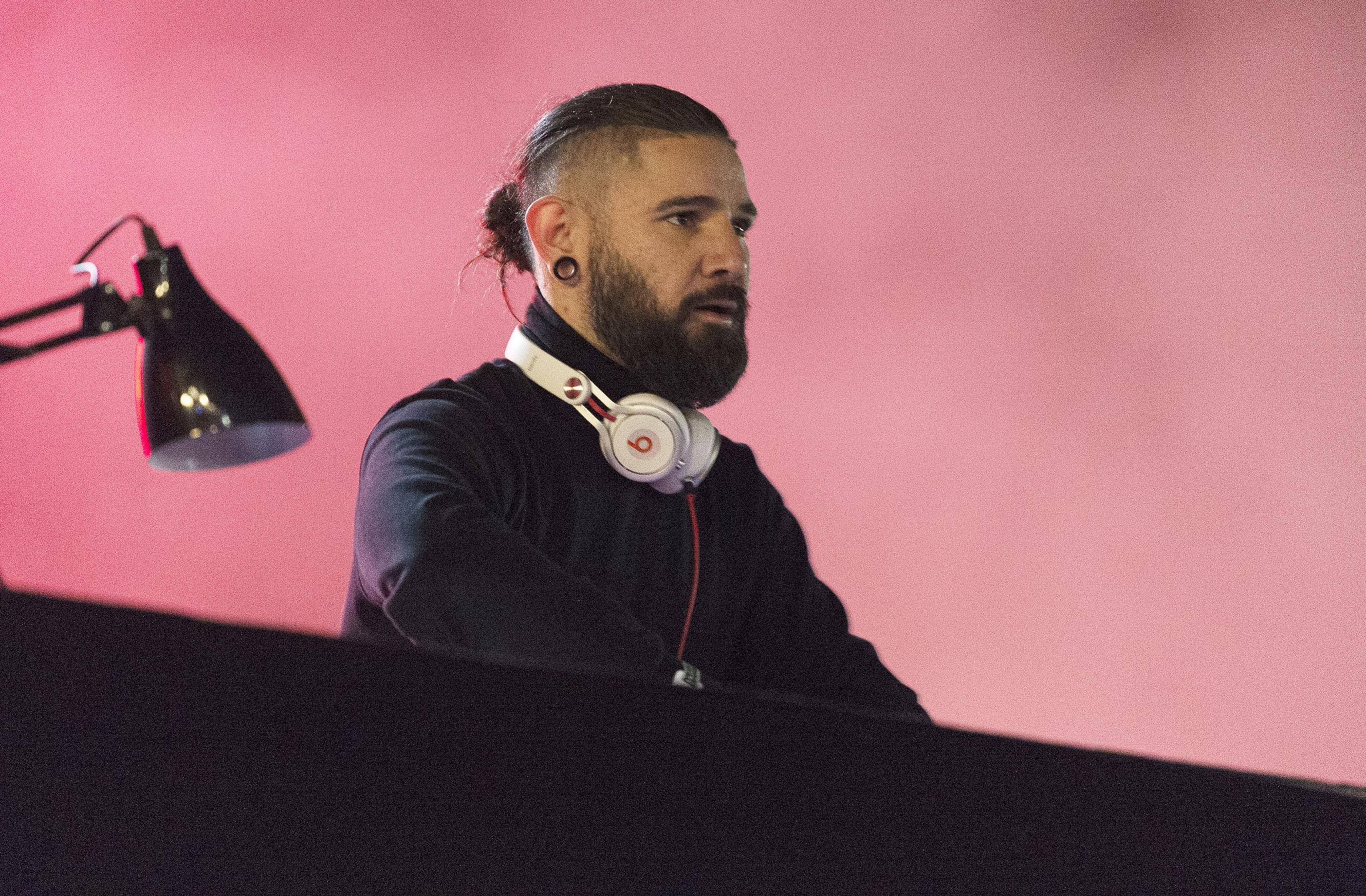A man with a beard and a top knot hairstyle stands behind a table, wearing white headphones. A desk lamp is on the left, and the background is a pinkish hue.