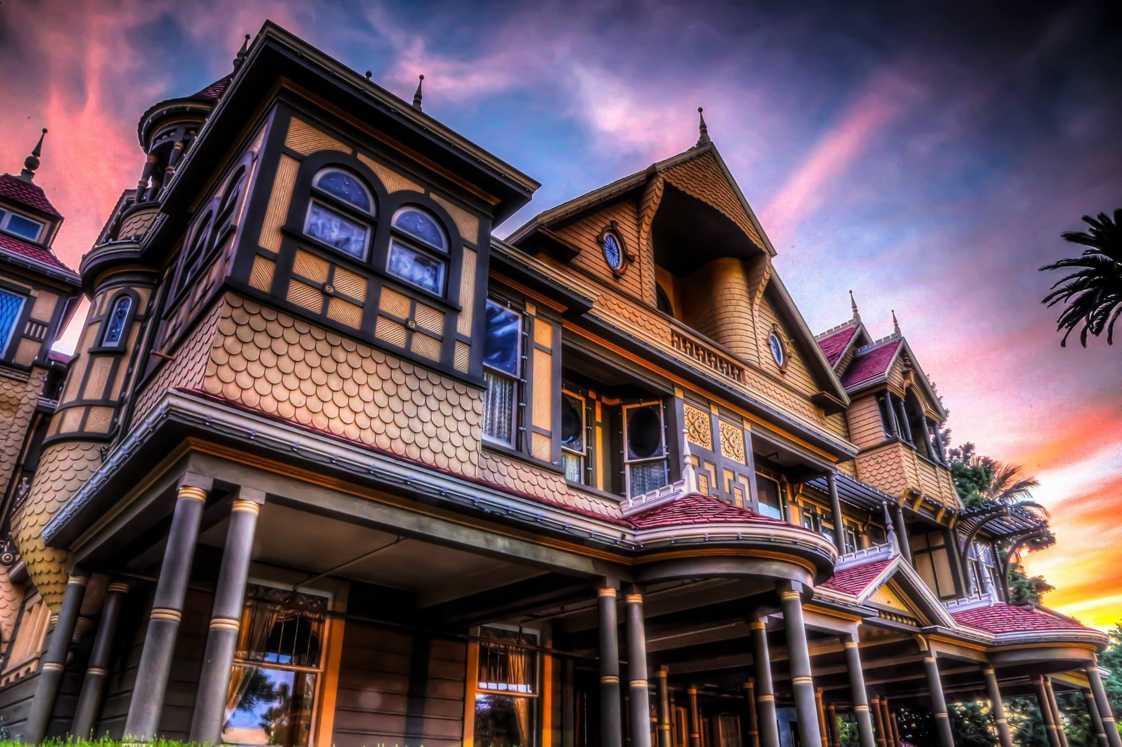 A large, ornate Victorian-style mansion with detailed woodwork, multiple windows, and tall columns under a vibrant pink and blue sunset sky.