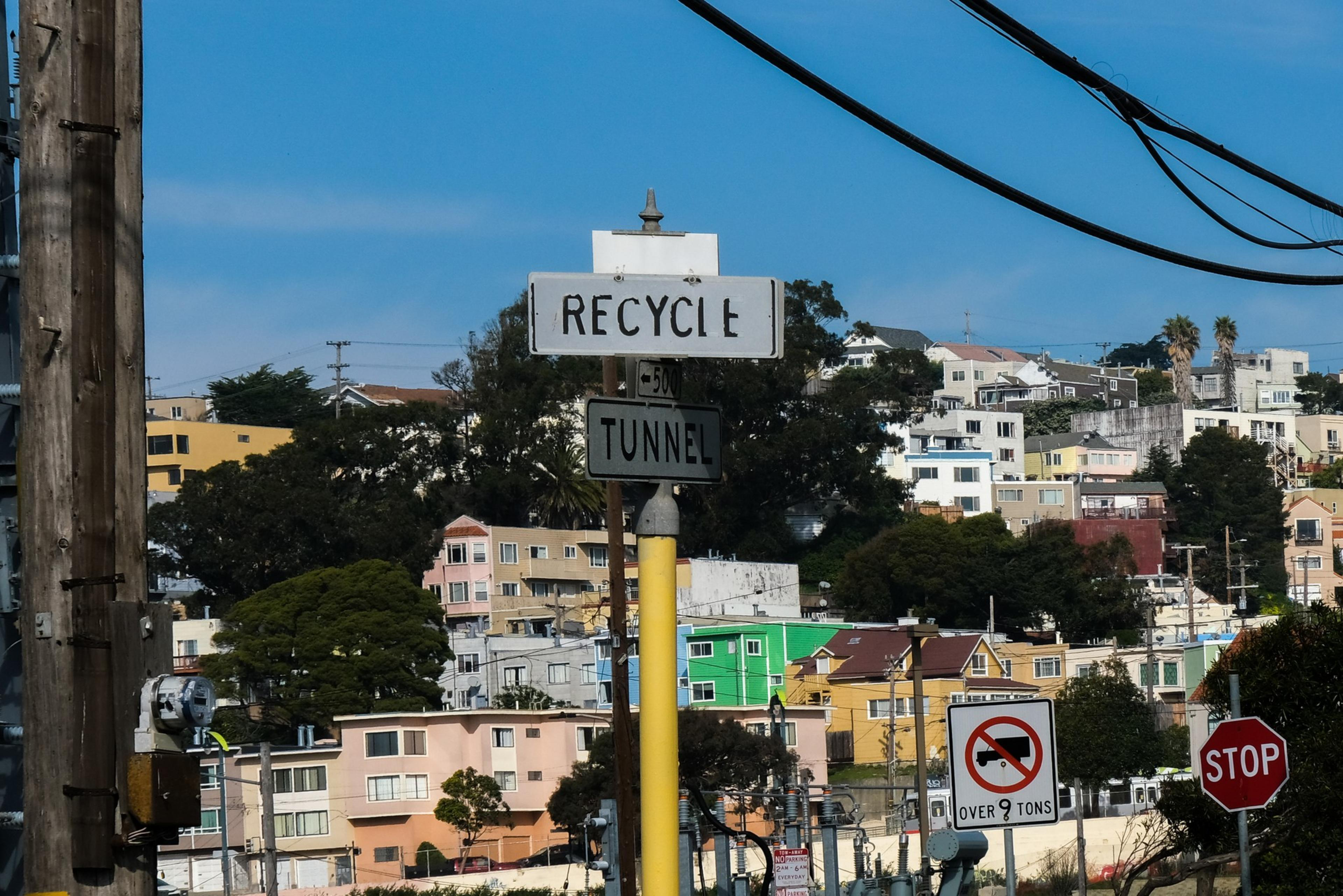 A street scene with a sign reading "RECYCLE" and "TUNNEL," colorful houses on a hillside, and a "No trucks over 9 tons" sign near a stop sign.