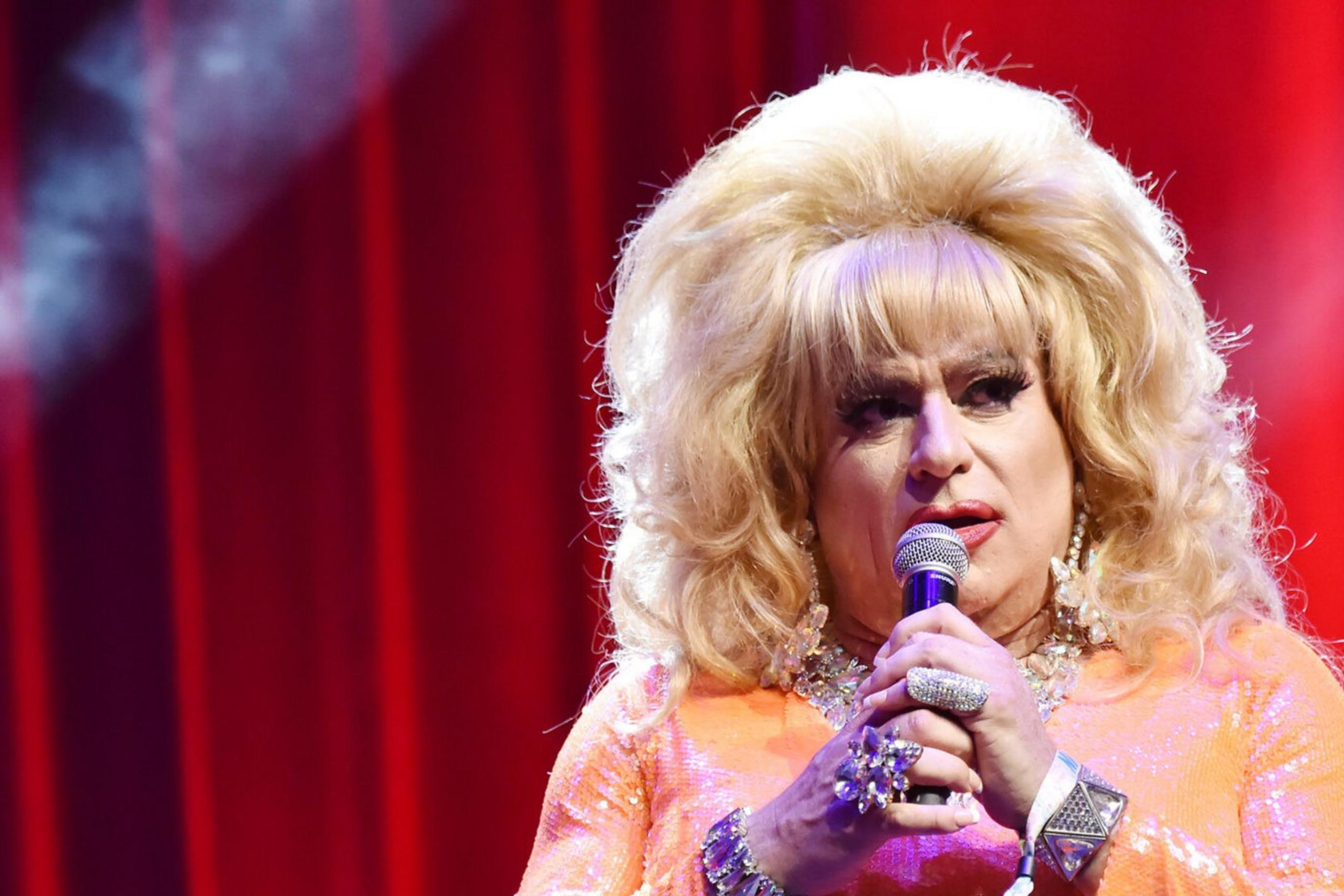 A performer with voluminous blonde hair and an orange sequined dress is holding a microphone on stage, set against a vibrant red curtain backdrop.
