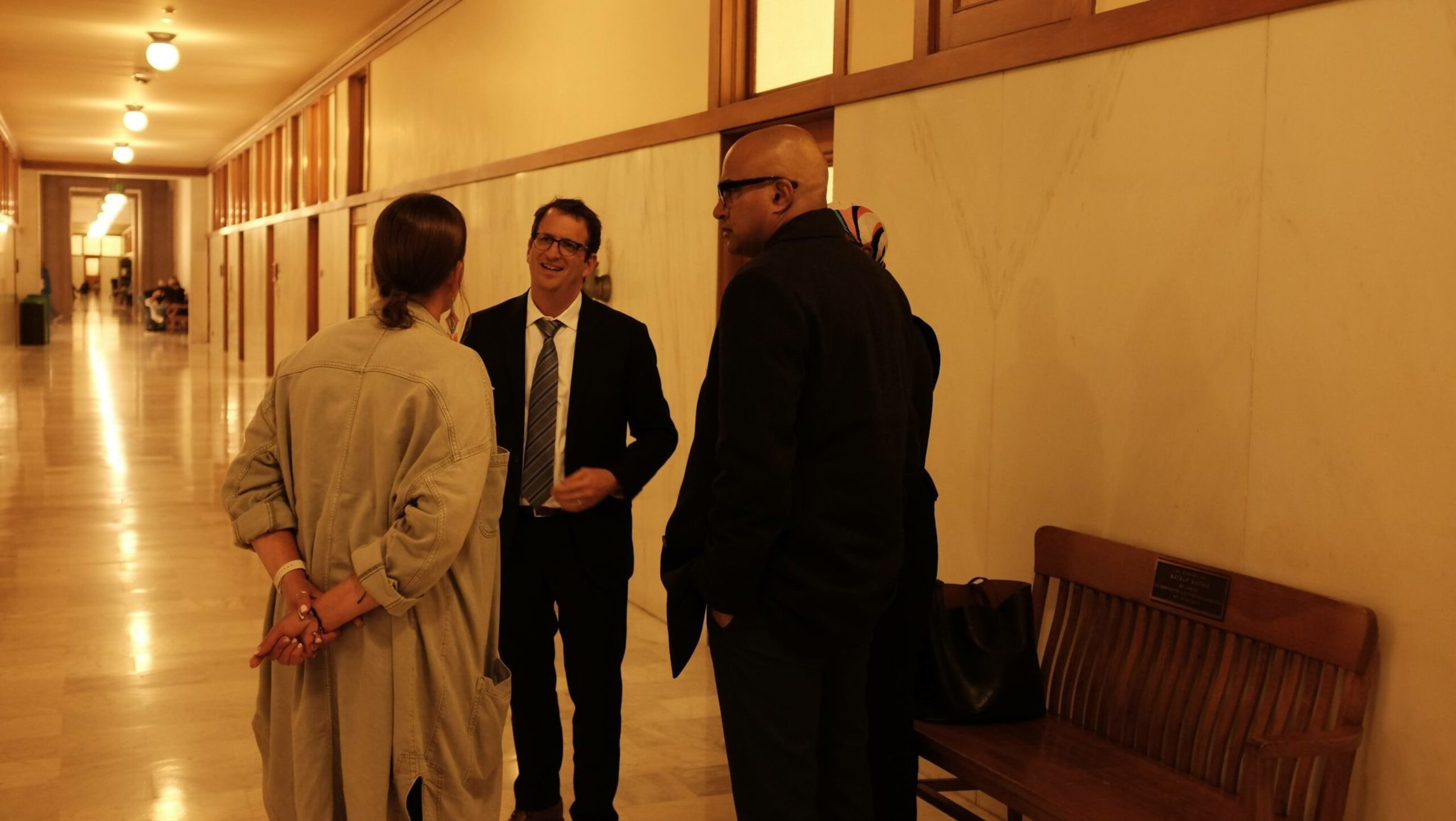 Four people, three men and one woman, engage in conversation in a well-lit hallway with marble walls and a wooden bench.