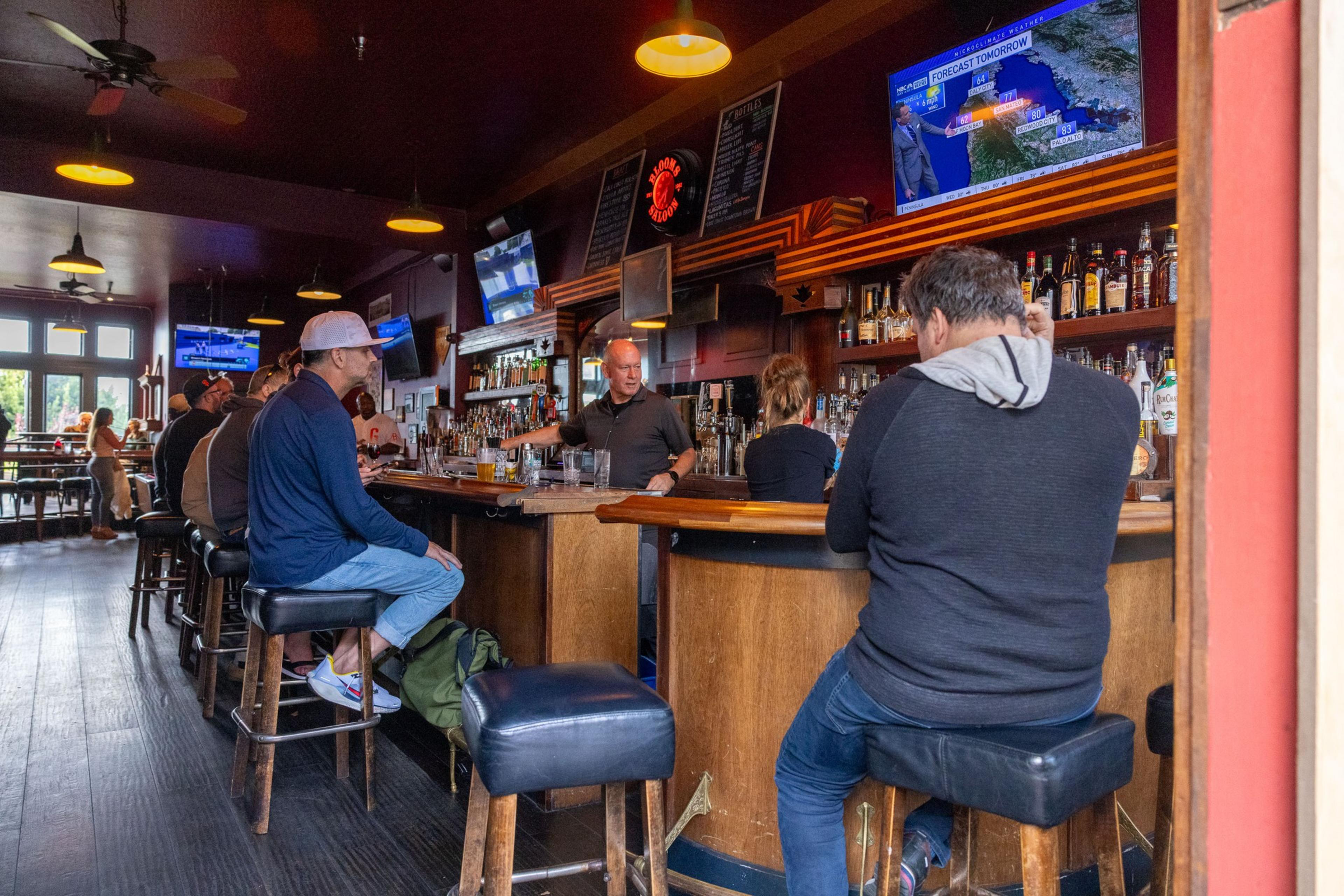 People sit at a bar counter inside a dimly lit pub, watching TV and conversing; bottles and glasses are visible on the shelves, with staff serving drinks.