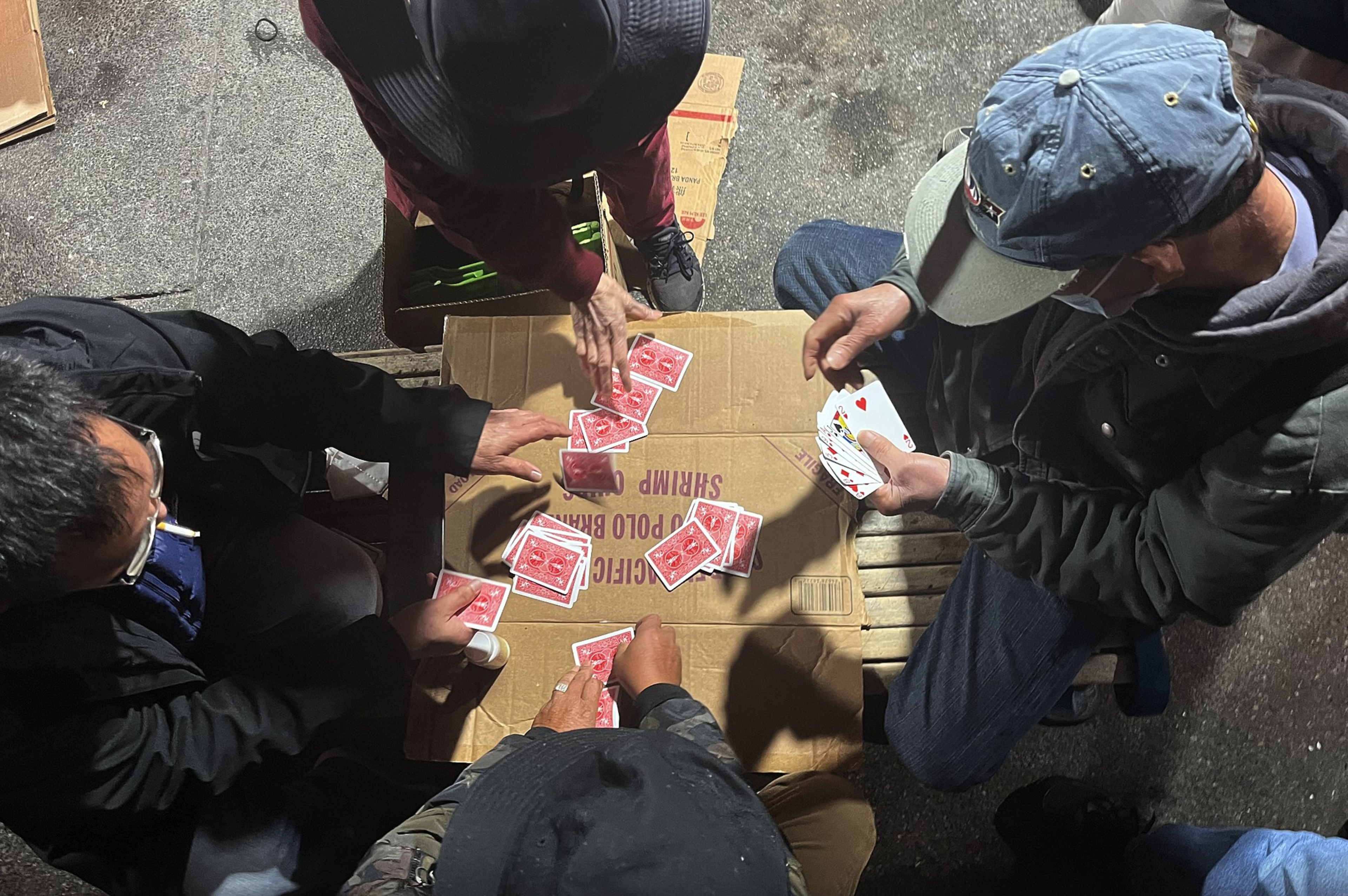Four people sit around a cardboard surface on the ground, playing cards, with some cards faced down and others held in their hands.