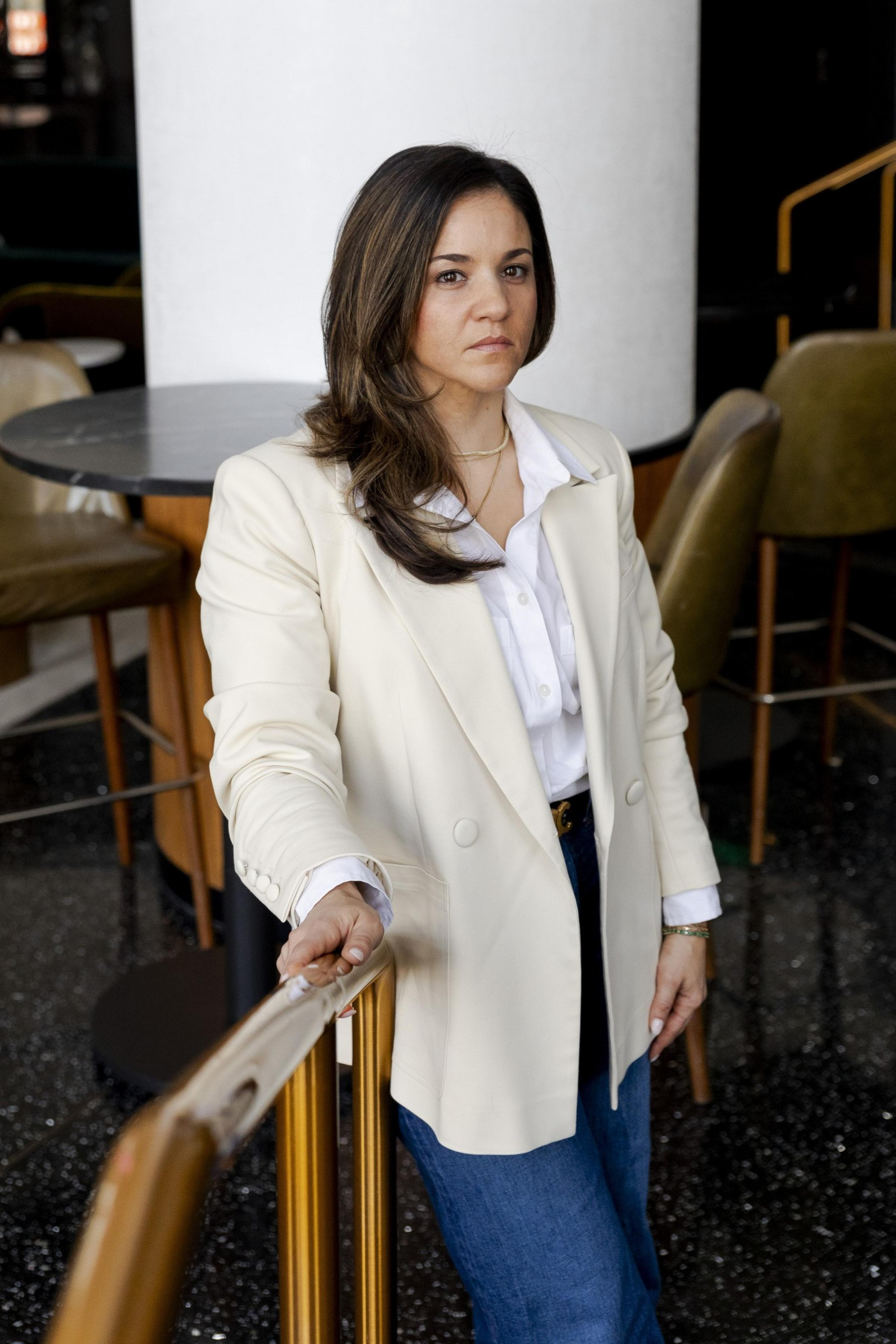 A woman in a white blazer and jeans stands indoors, holding a railing. Behind her, there are brown chairs and a round table. She looks confidently toward the camera.