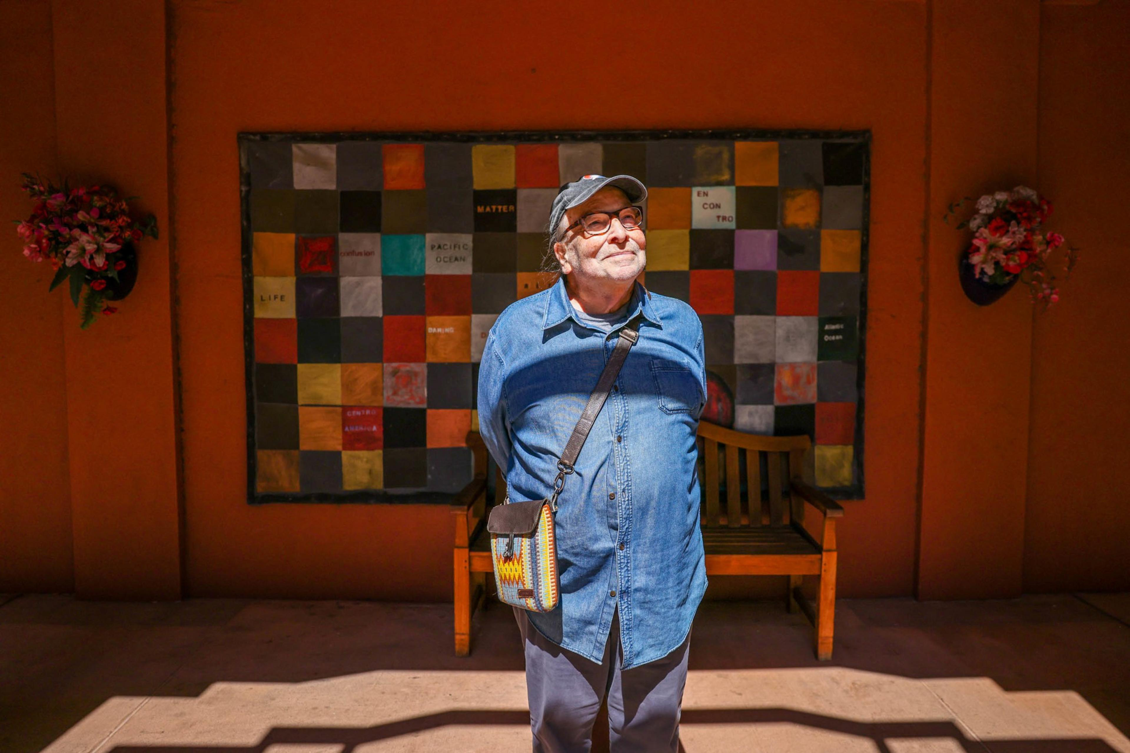 An older man wearing a blue shirt and cap stands with hands behind his back, smiling, in front of a colorful patchwork wall with two flower pots.