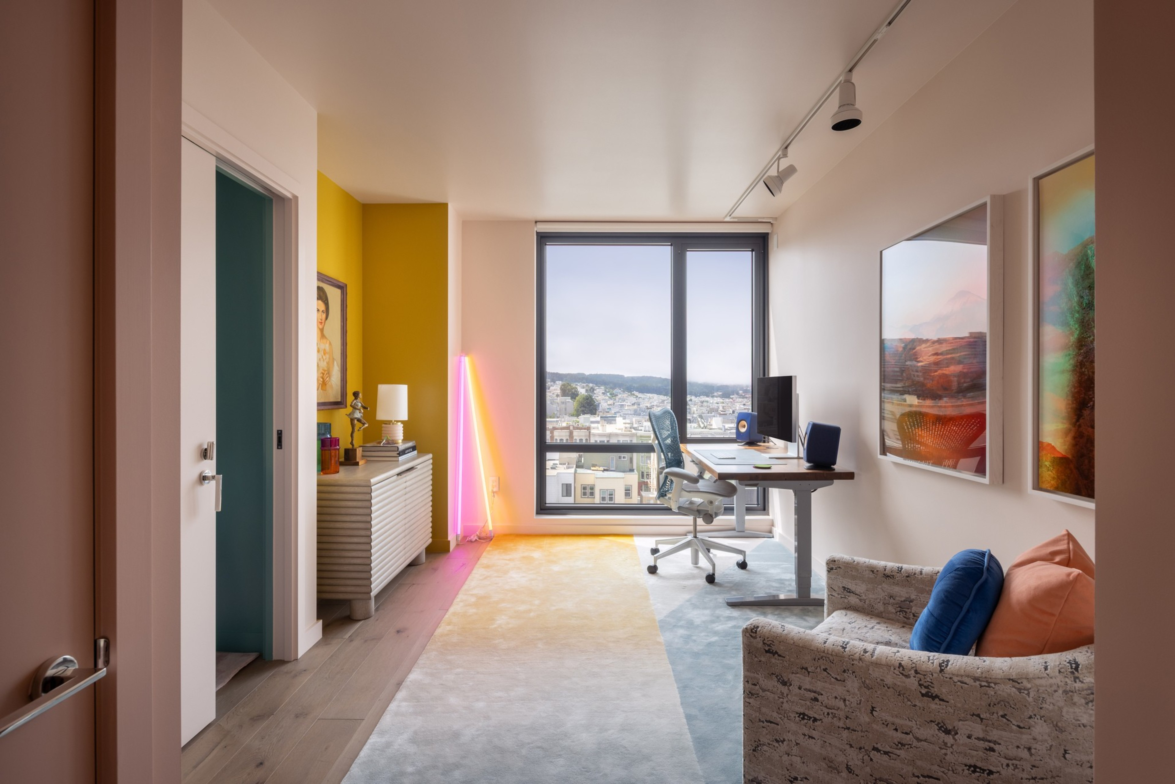 A modern home office features a large window with a city view, a desk with a computer, a patterned chair with blue and orange pillows, and colorful wall art.