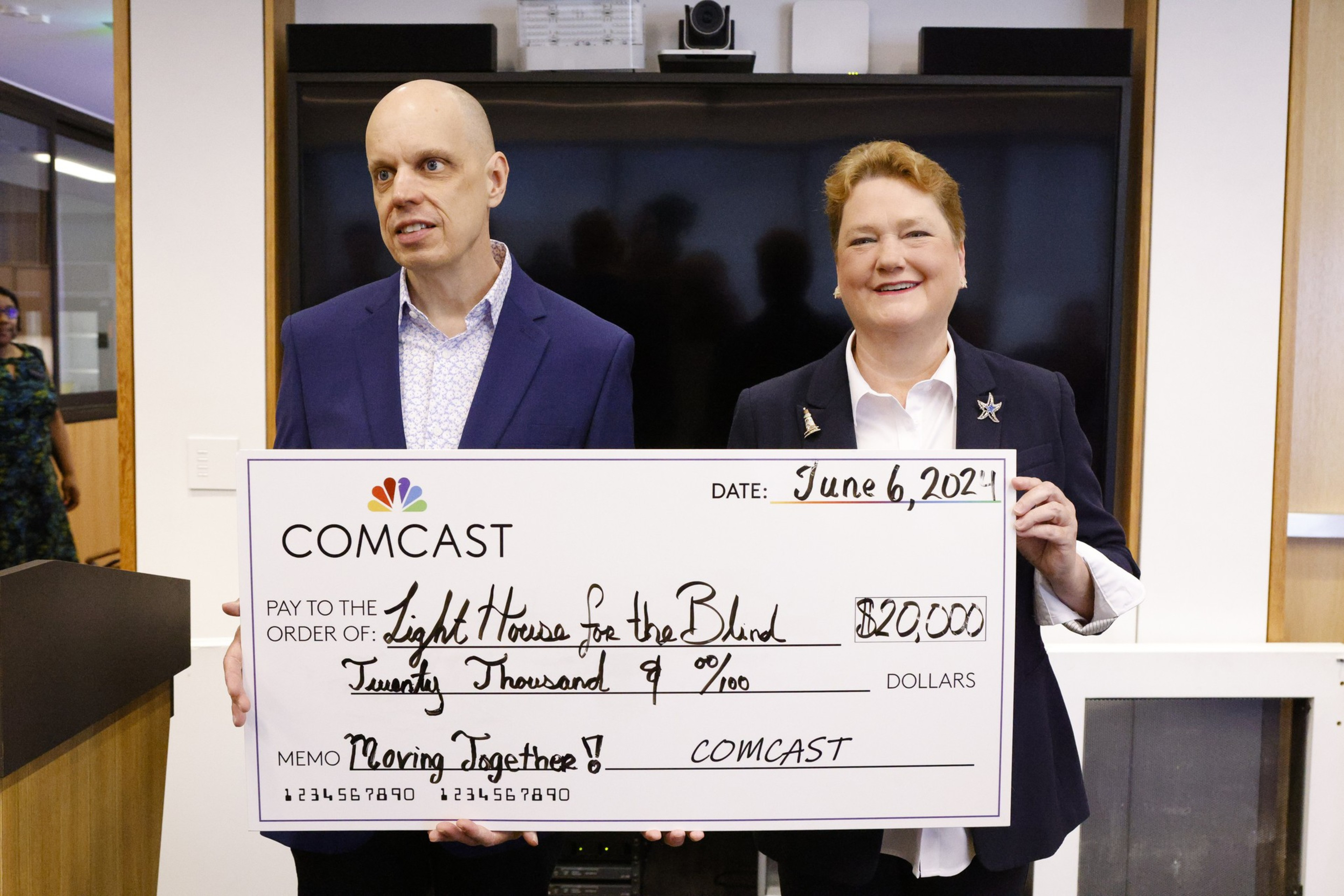 Two people in business attire are holding a giant $20,000 check from Comcast addressed to &quot;Lighthouse for the Blind.&quot;