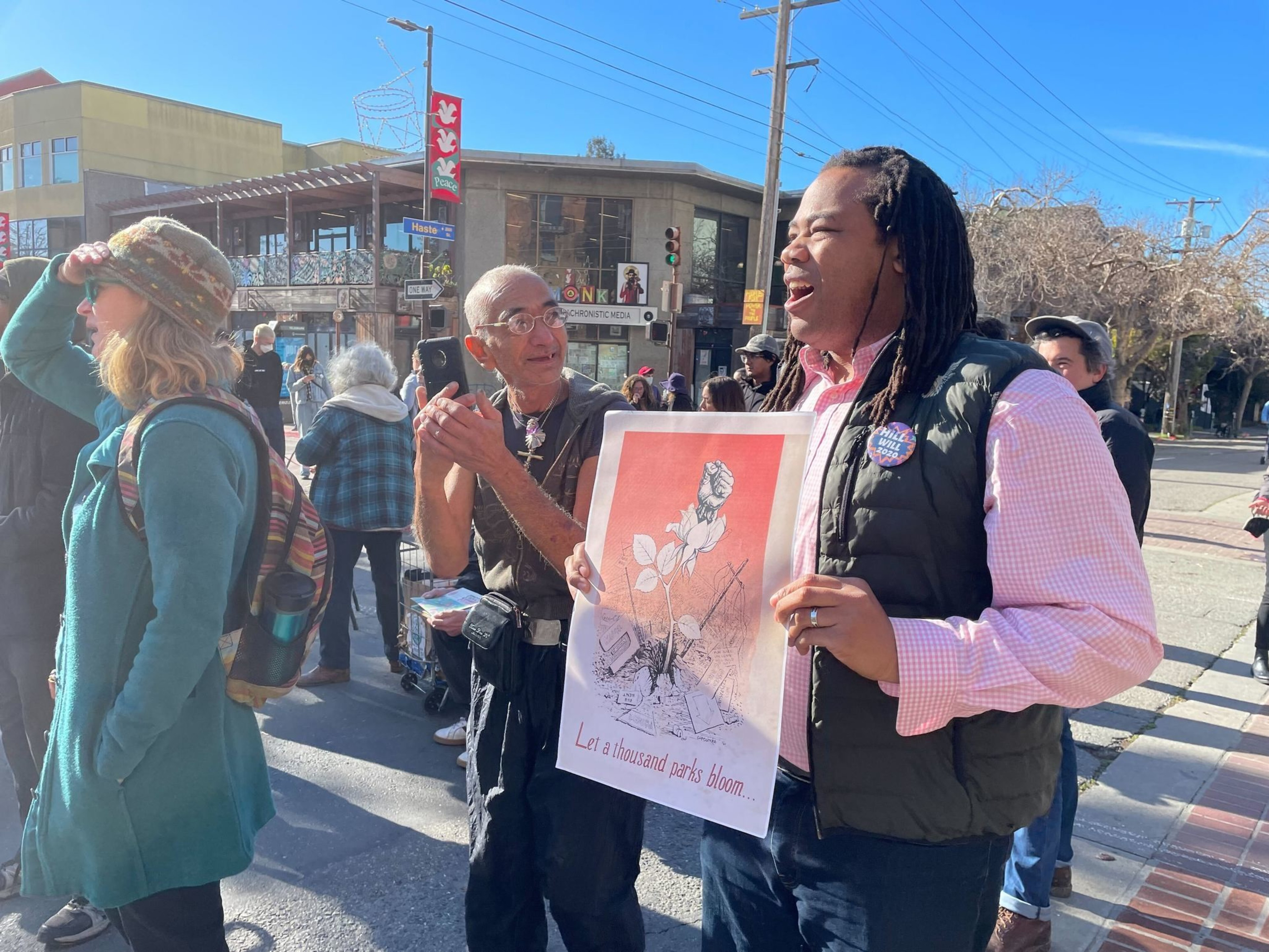 A person holds a sign and chants next to other people standing near a city intersection.