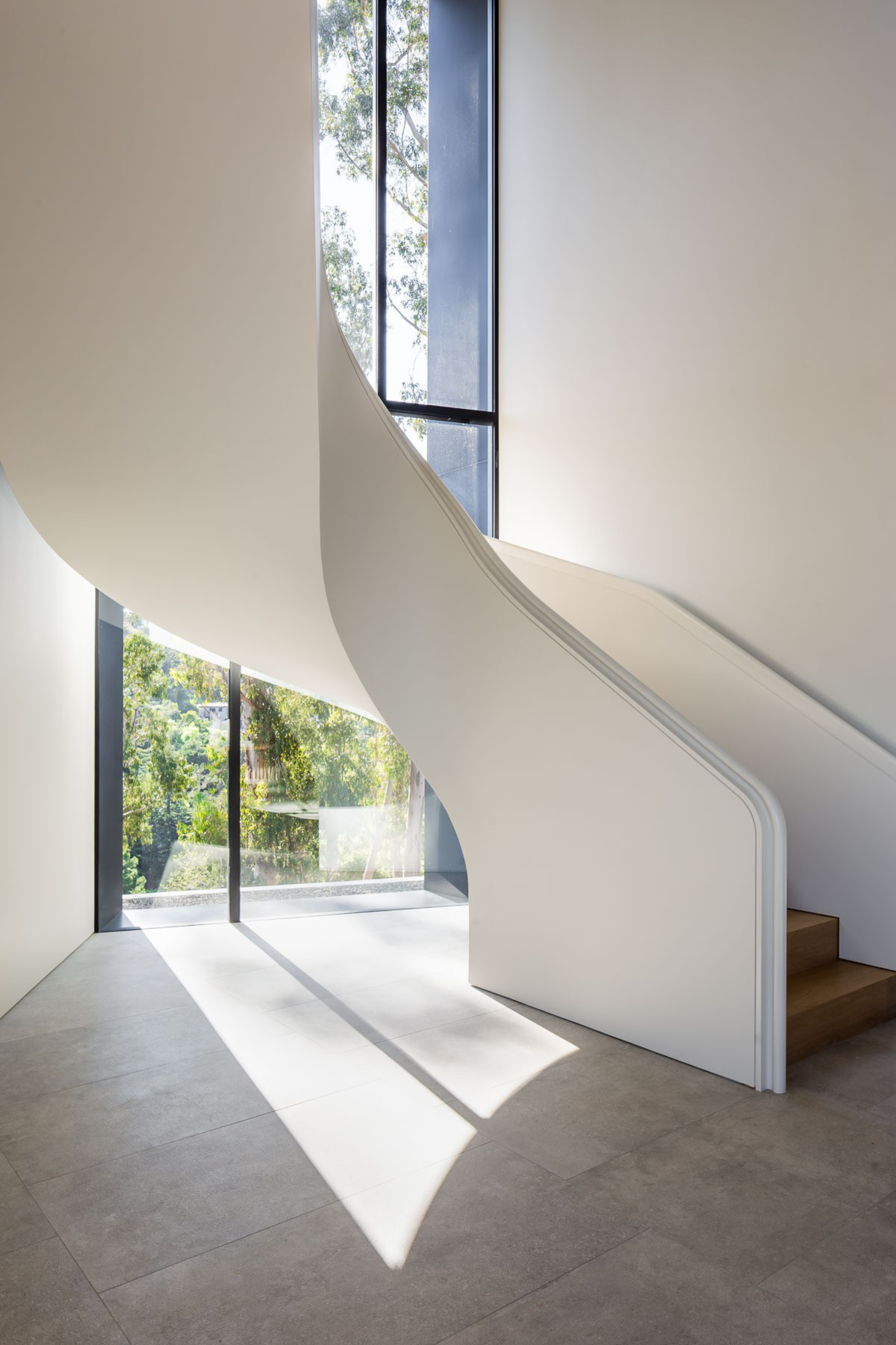 A sleek, white spiral staircase curves upward next to tall windows, casting soft natural light and shadows on the light-tiled floor below.