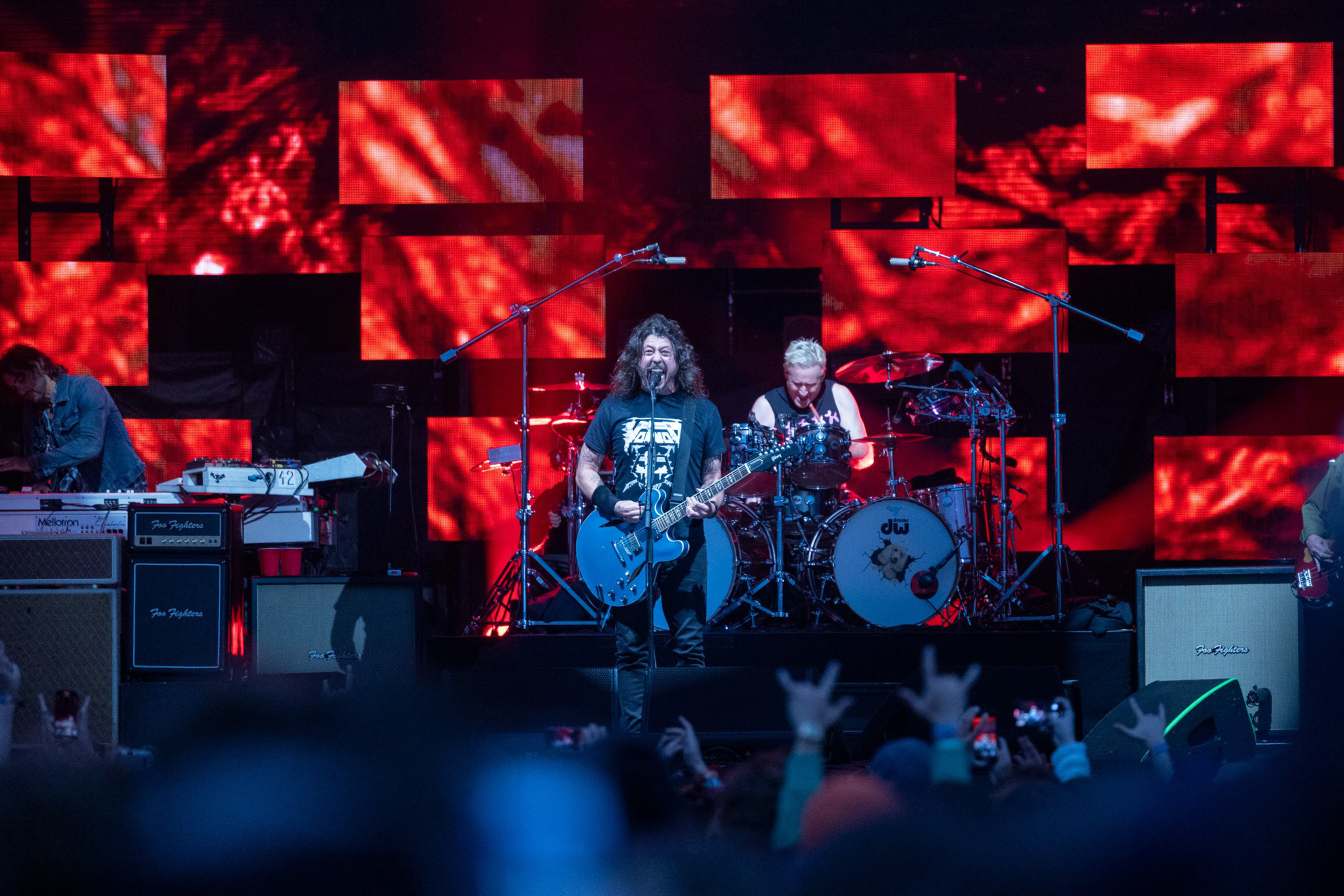 A rock band performs on stage with a singer playing a blue guitar and a drummer behind, as red abstract visuals light up the background.