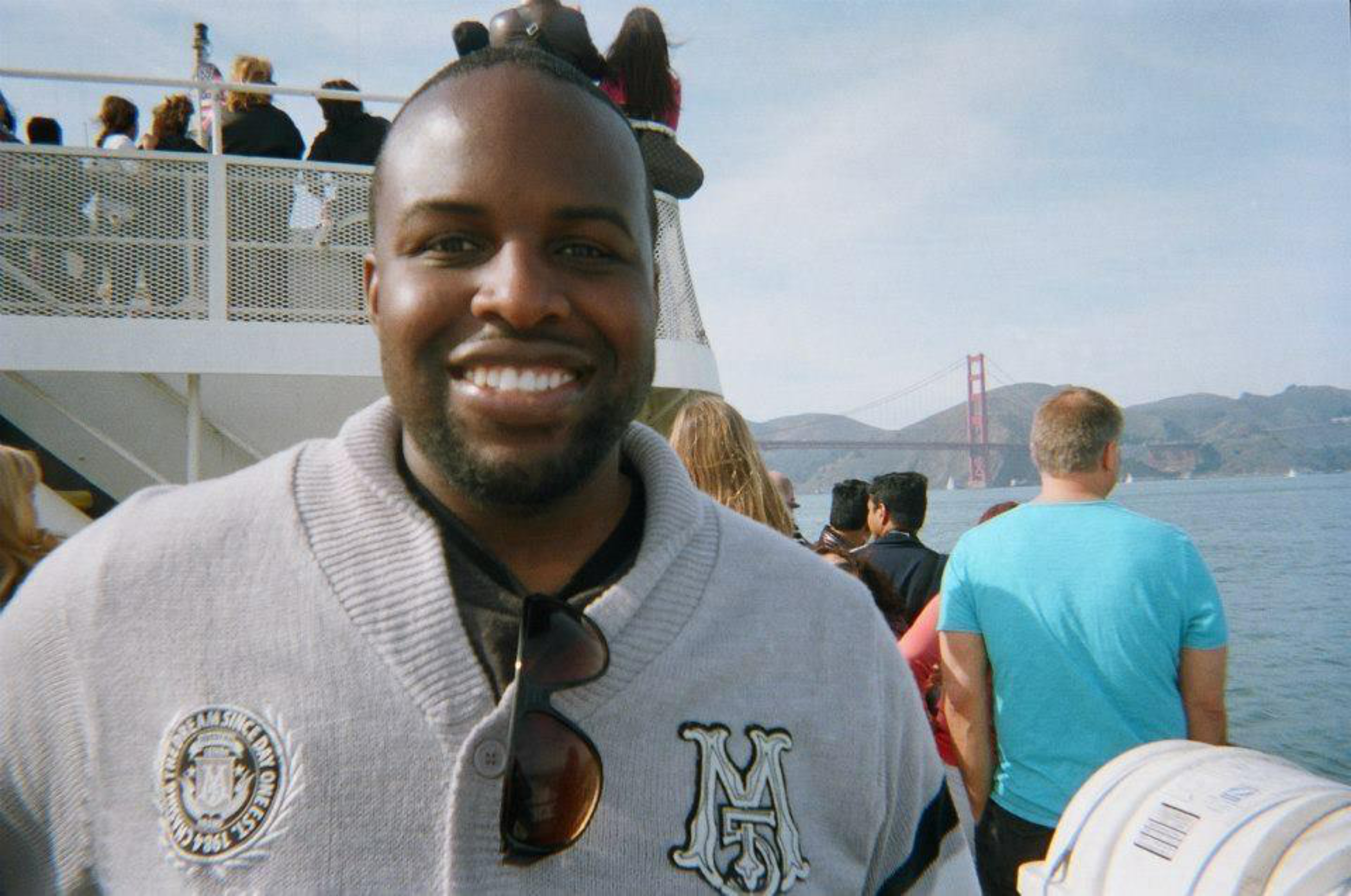 A smiling man wearing a gray sweater with sunglasses stands on a boat with the Golden Gate Bridge and other passengers in the background.