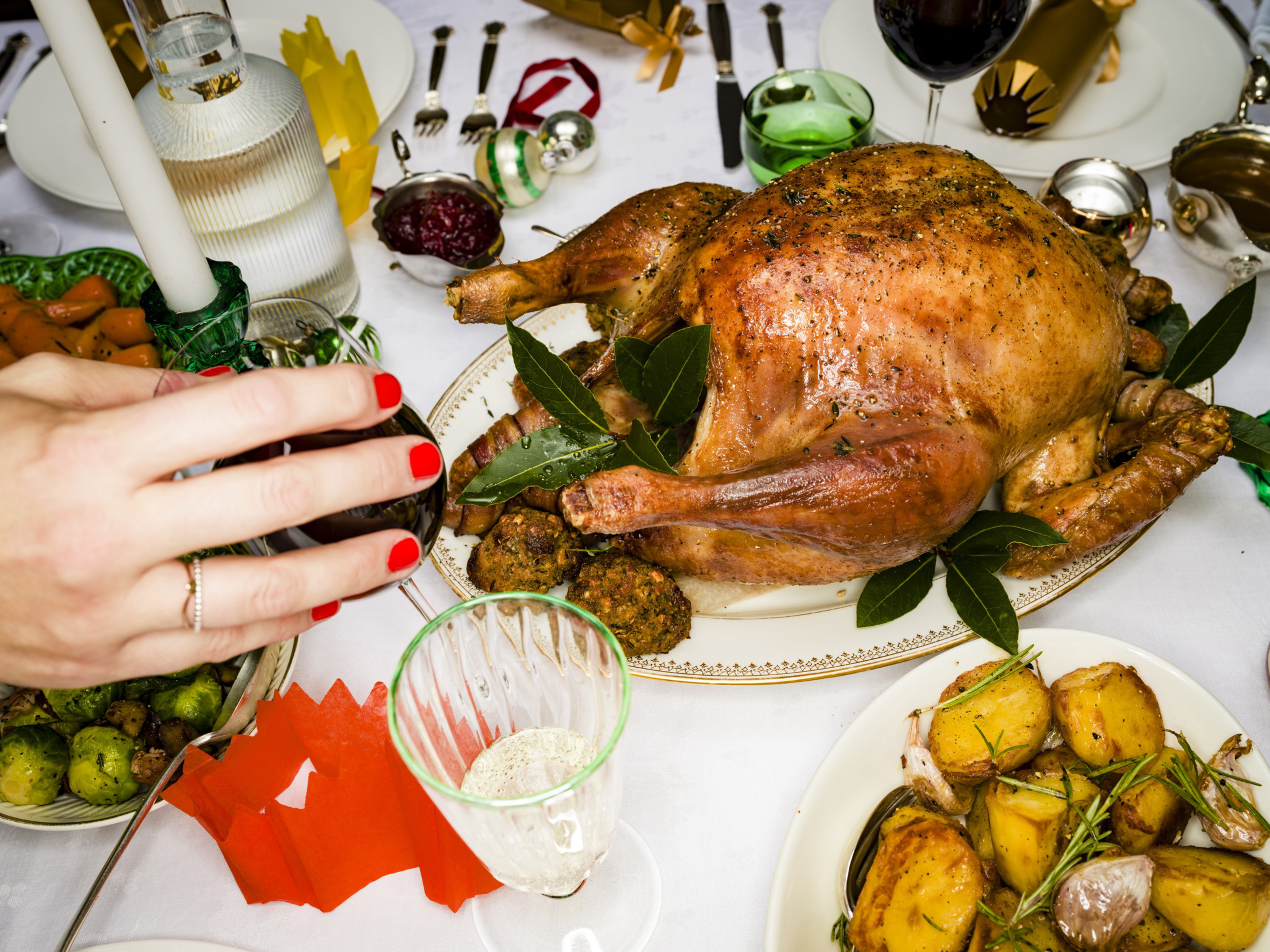 A golden roasted turkey garnished with green leaves and stuffing sits on a platter surrounded by roasted potatoes, vegetables, wine, and holiday decorations.