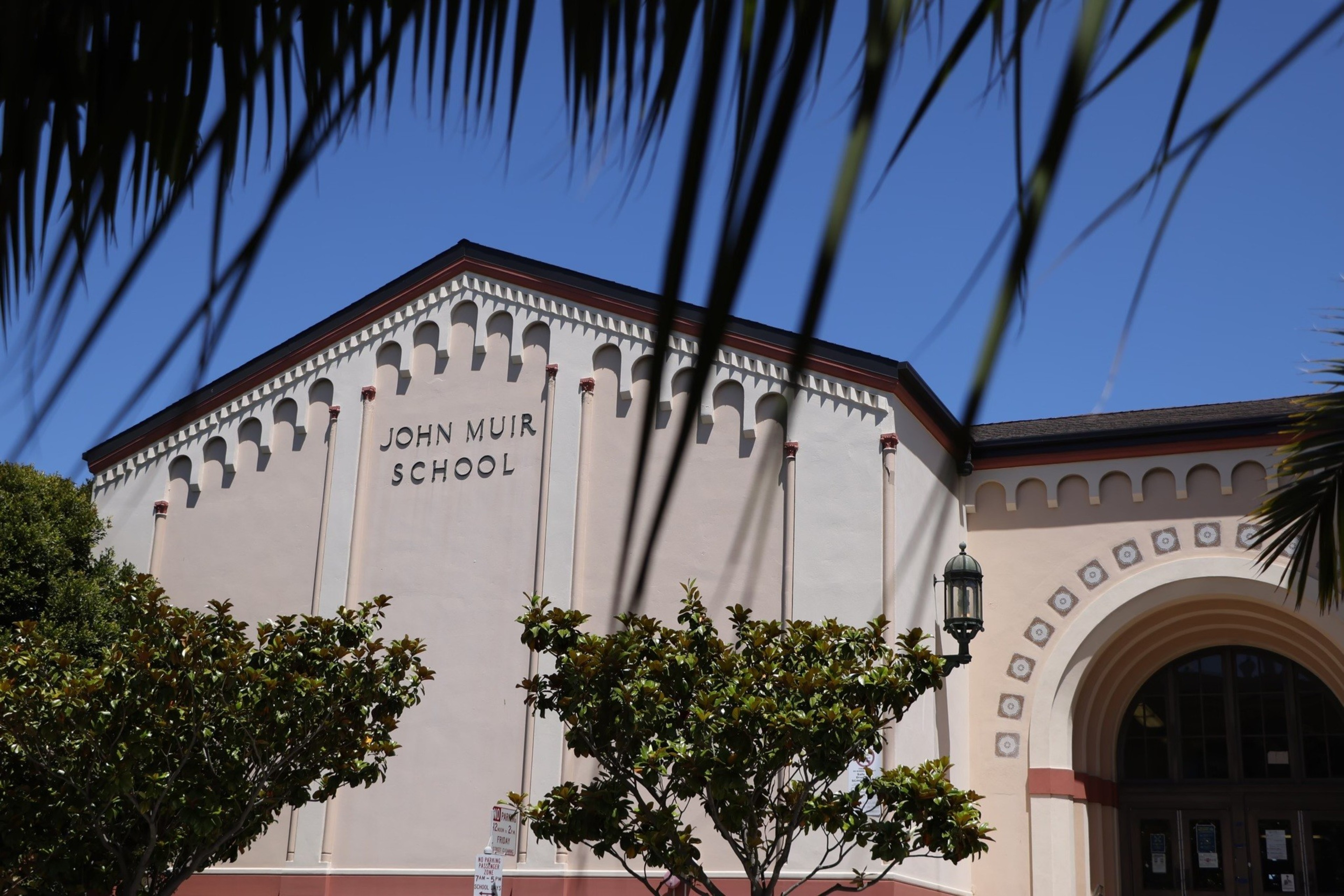 A beige school building with "John Muir School" written on it, framed by tree branches and a clear blue sky.