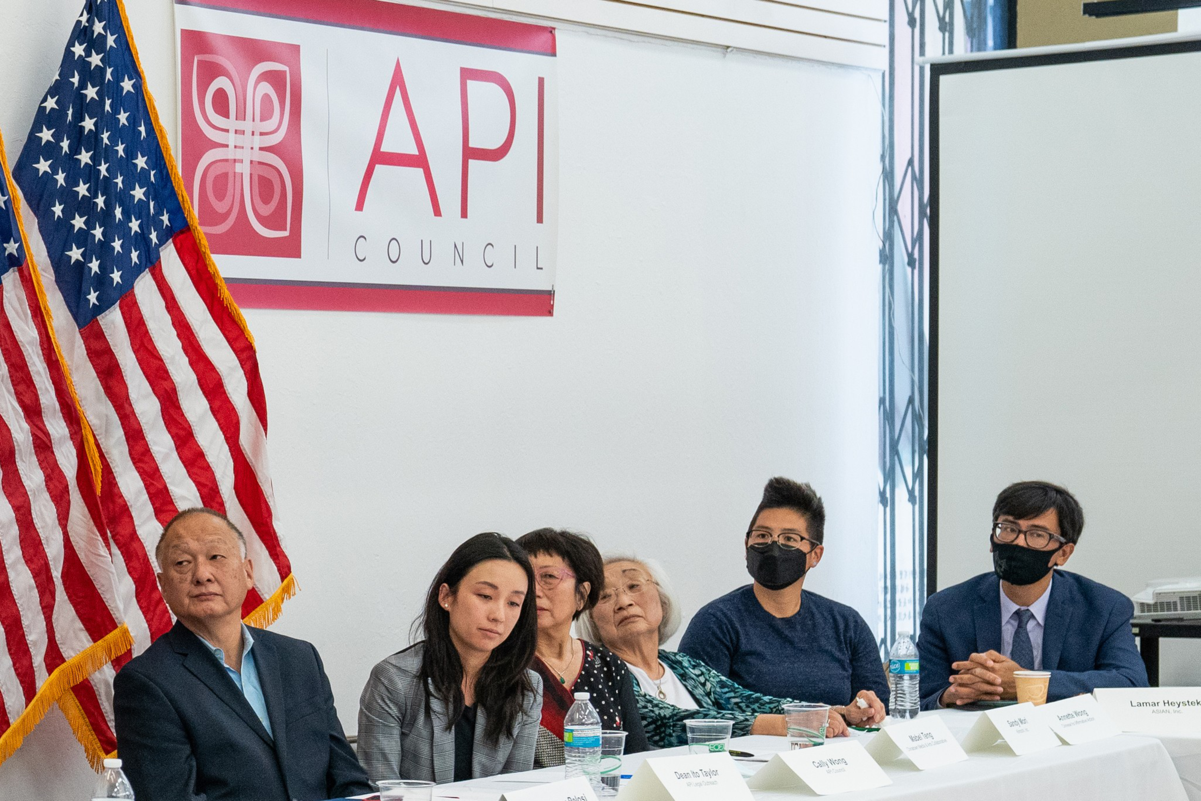 Six people sit at a table with nameplates, an American flag stands behind them, and a banner reading "API Council" hangs on the wall.