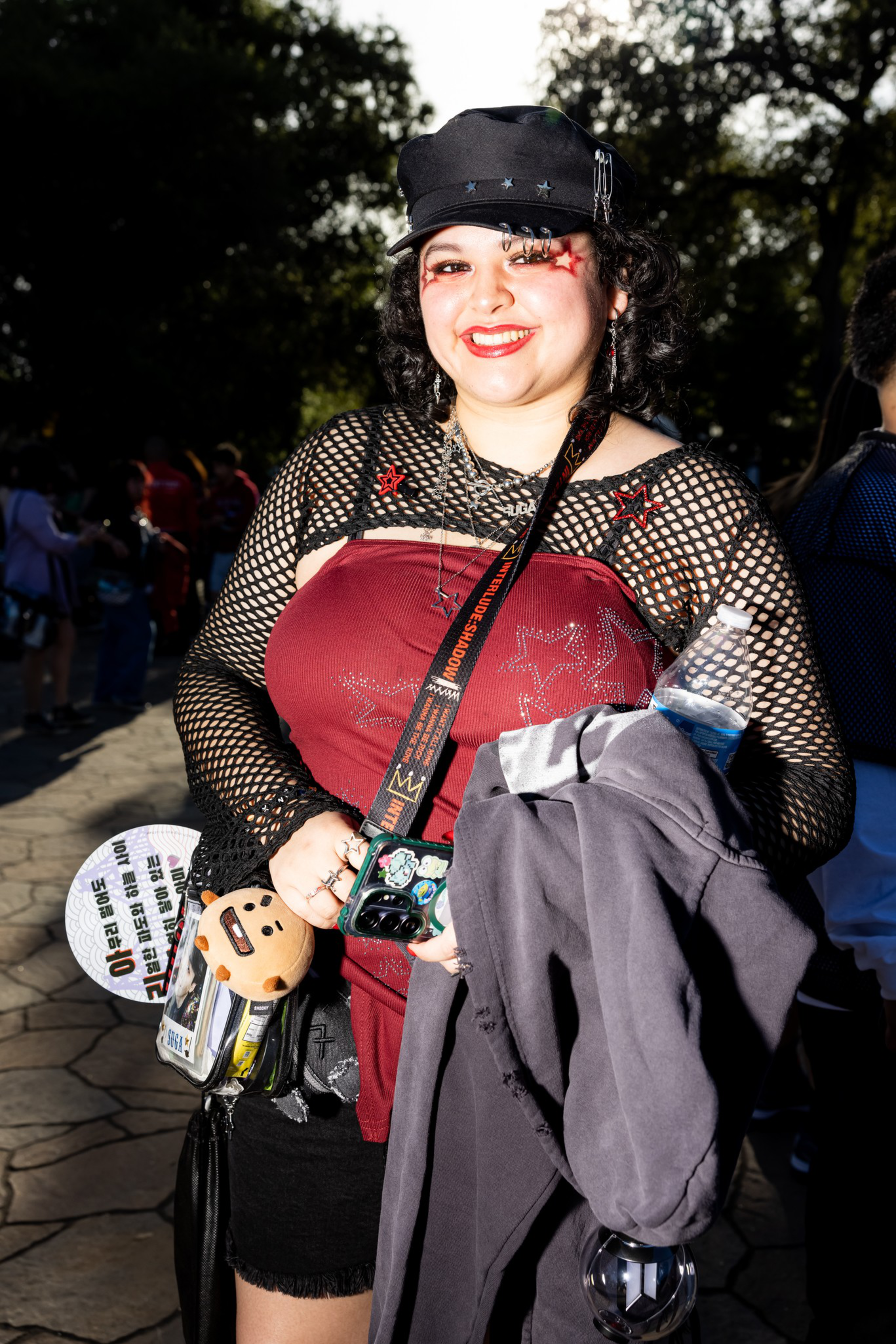 A smiling person wears a black cap, fishnet top over a red star-patterned dress, holds a jacket, water bottle, and phone with a cartoon keychain.