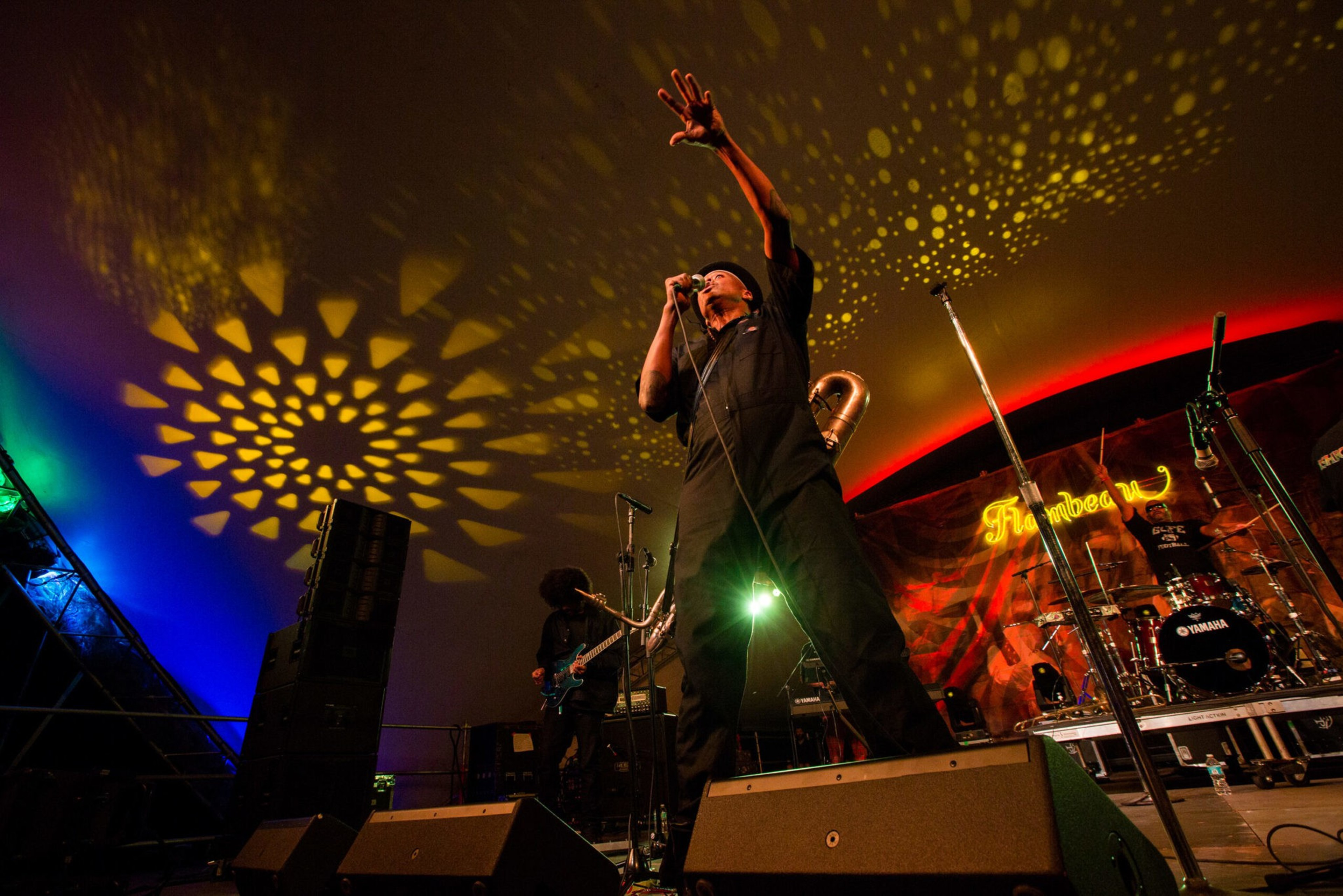 A musician sings passionately into a microphone with one arm raised, colorful stage lights casting patterned shadows on the ceiling behind him.