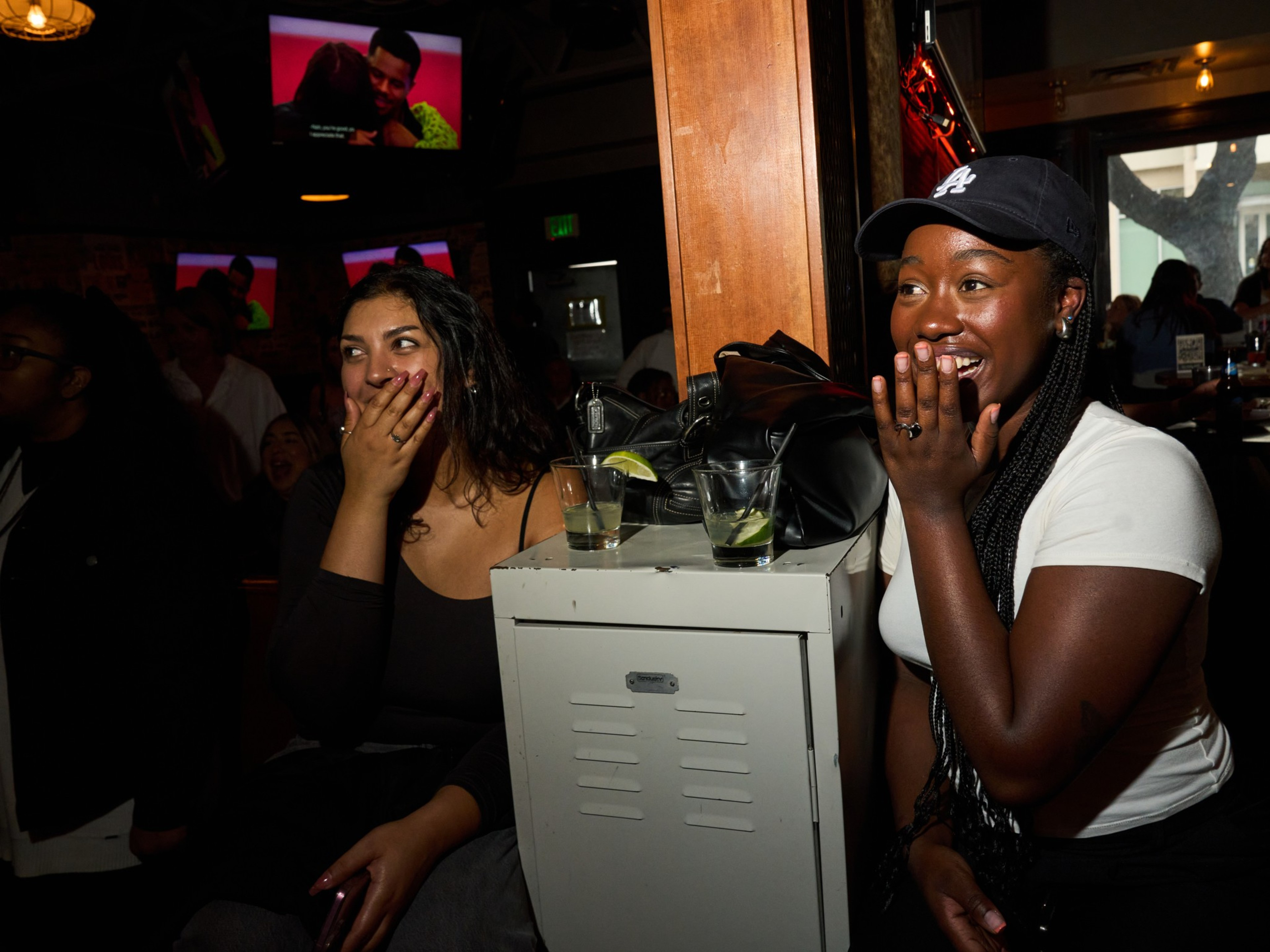 Two women are seated, watching a TV screen and covering their mouths in surprise or delight. They're in a dimly lit room with drinks and bags nearby.