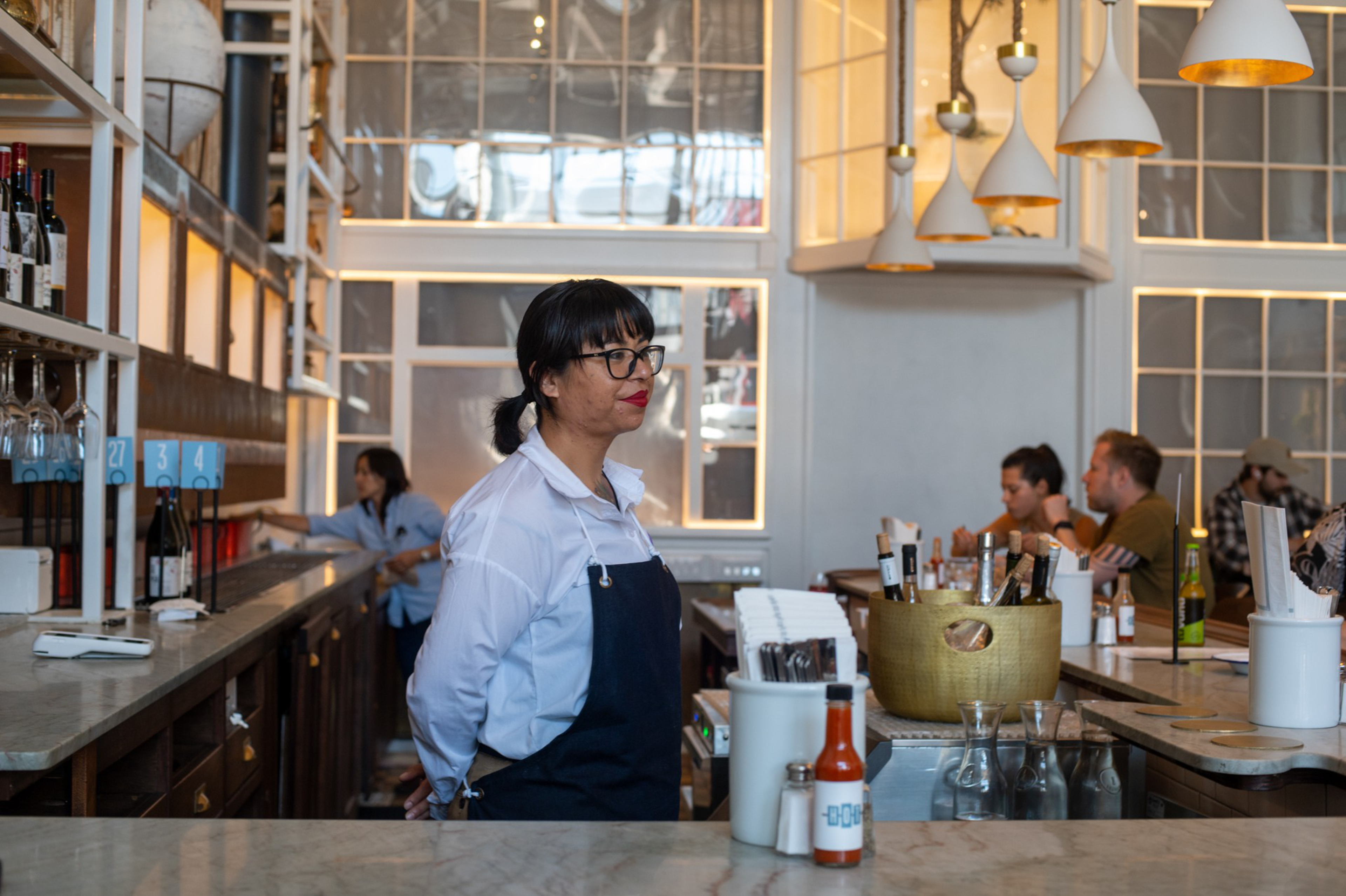 A woman wearing glasses, a white shirt, and a dark apron stands behind a bar counter, with other people seated at tables in a modern café.
