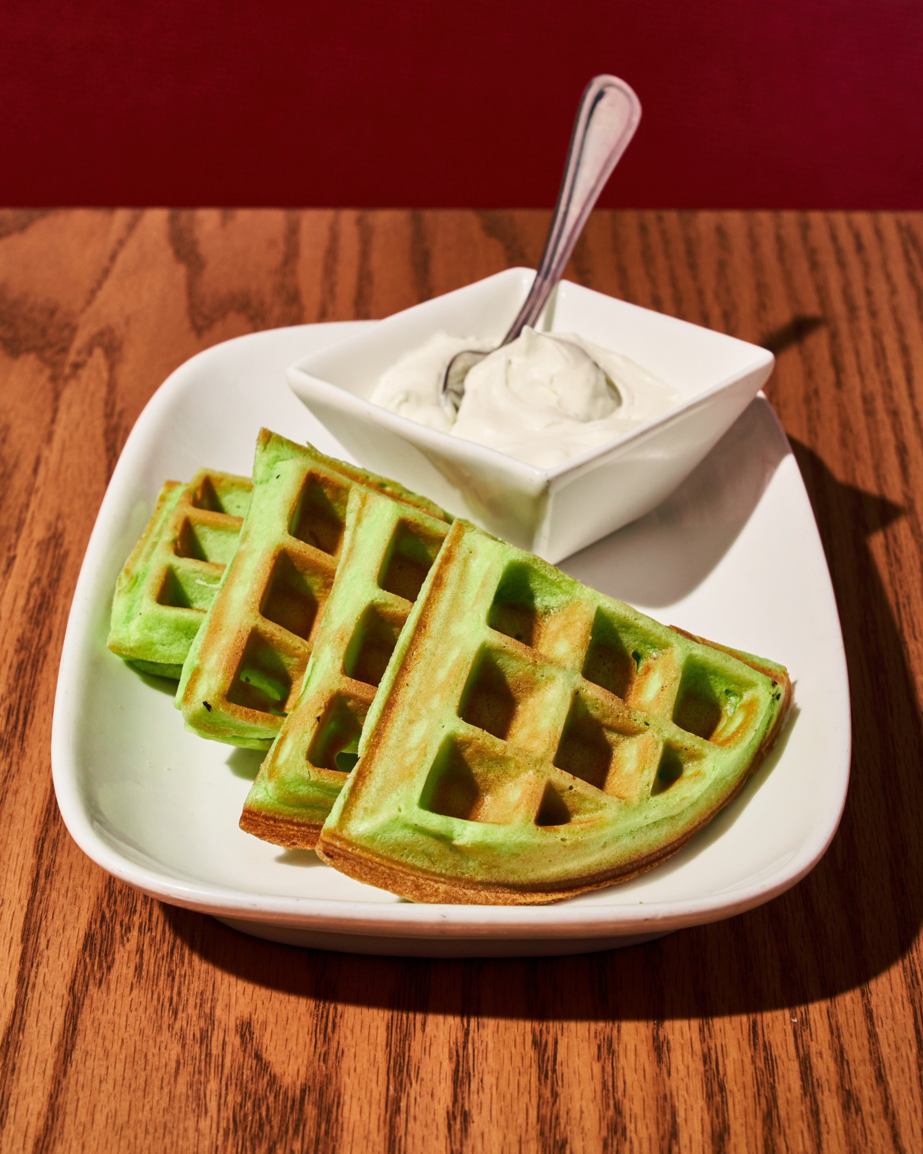 The image shows three pieces of green waffles on a white plate, accompanied by a square bowl of white cream with a spoon, set on a wooden table.