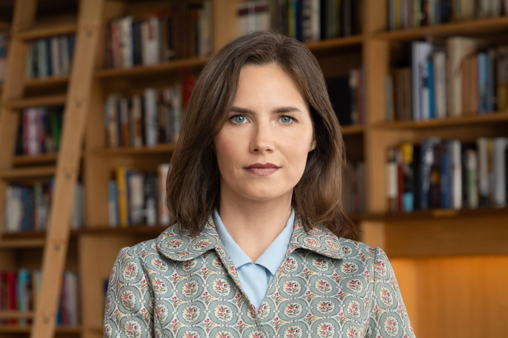 A woman with shoulder-length brown hair, wearing a patterned jacket over a light blue shirt and dark purple pants, stands in front of a bookshelf.