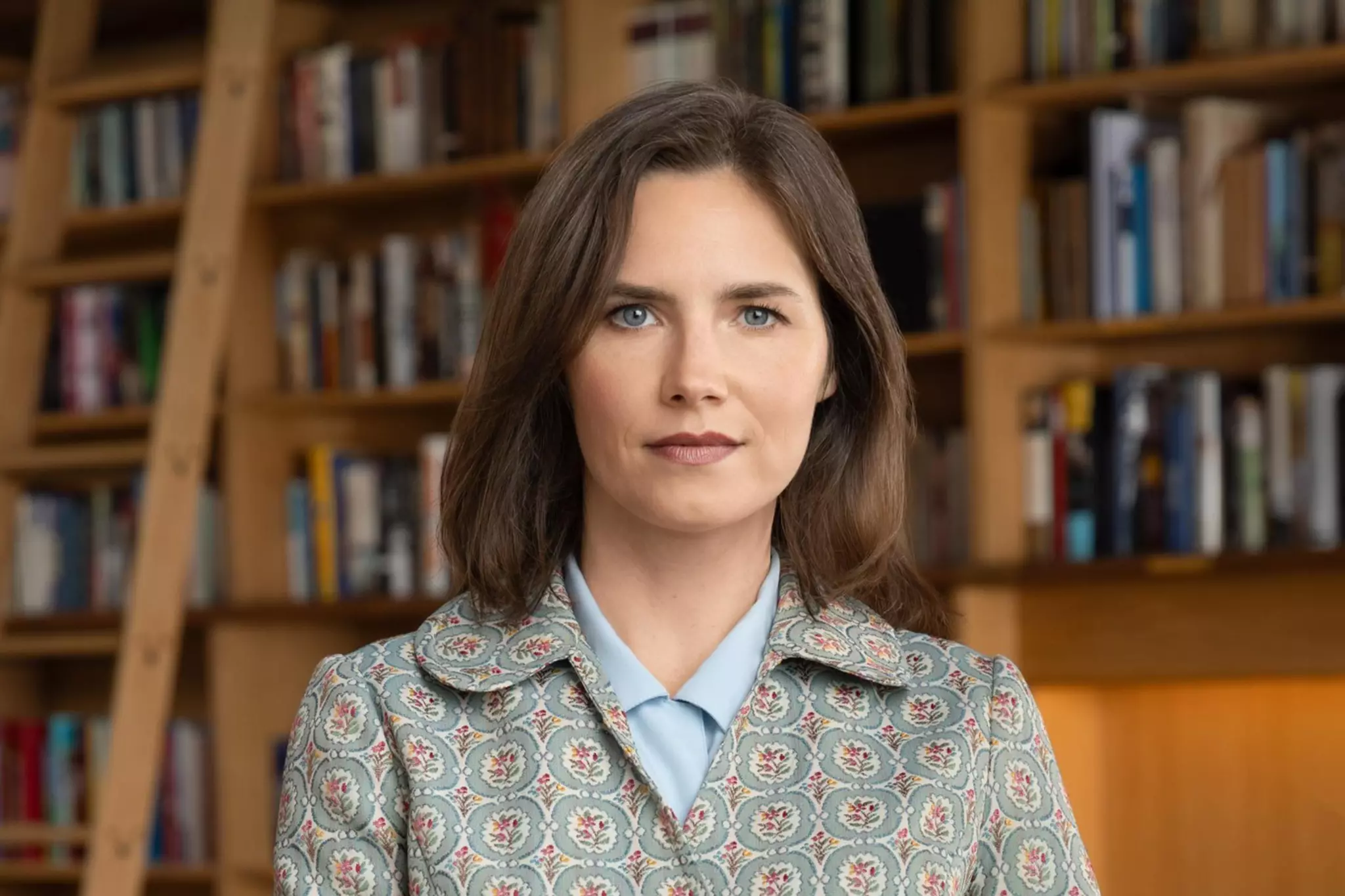 A woman with shoulder-length brown hair, wearing a patterned jacket over a light blue shirt and dark purple pants, stands in front of a bookshelf.