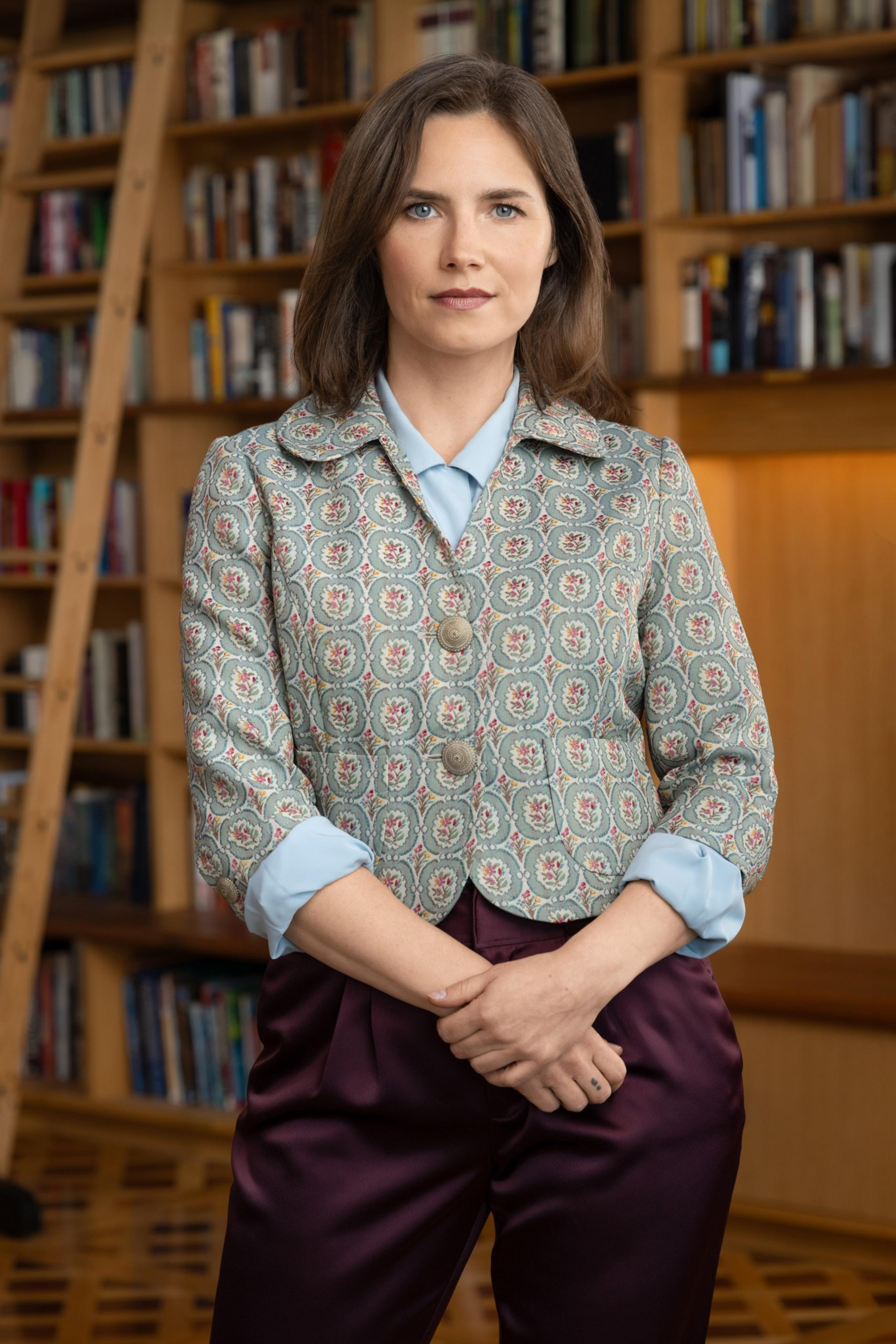 A woman with shoulder-length brown hair, wearing a patterned jacket over a light blue shirt and dark purple pants, stands in front of a bookshelf.