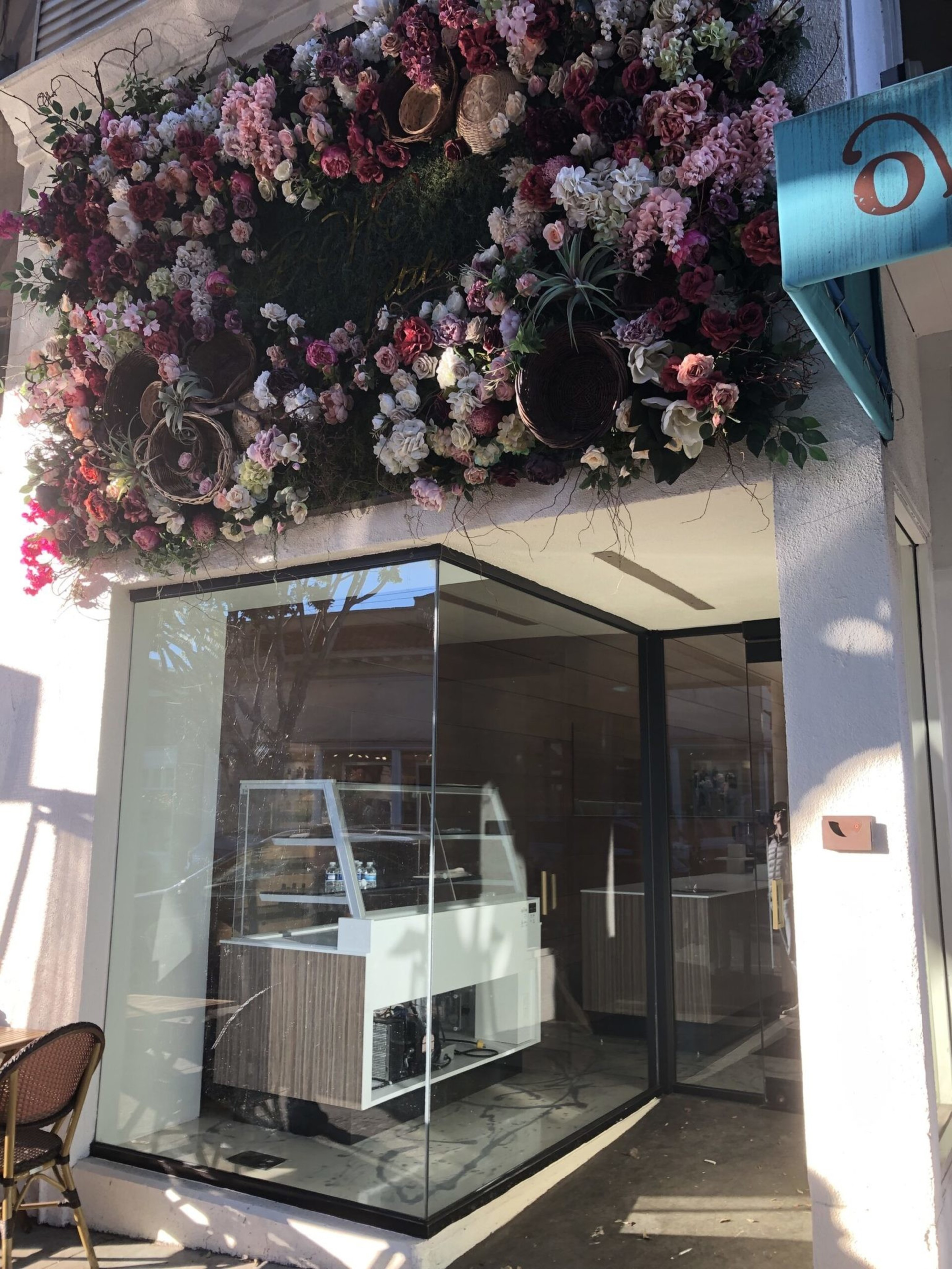 A storefront entrance with glass walls displays a counter inside, topped by a lush floral arrangement featuring various pink and white flowers above the doorway.