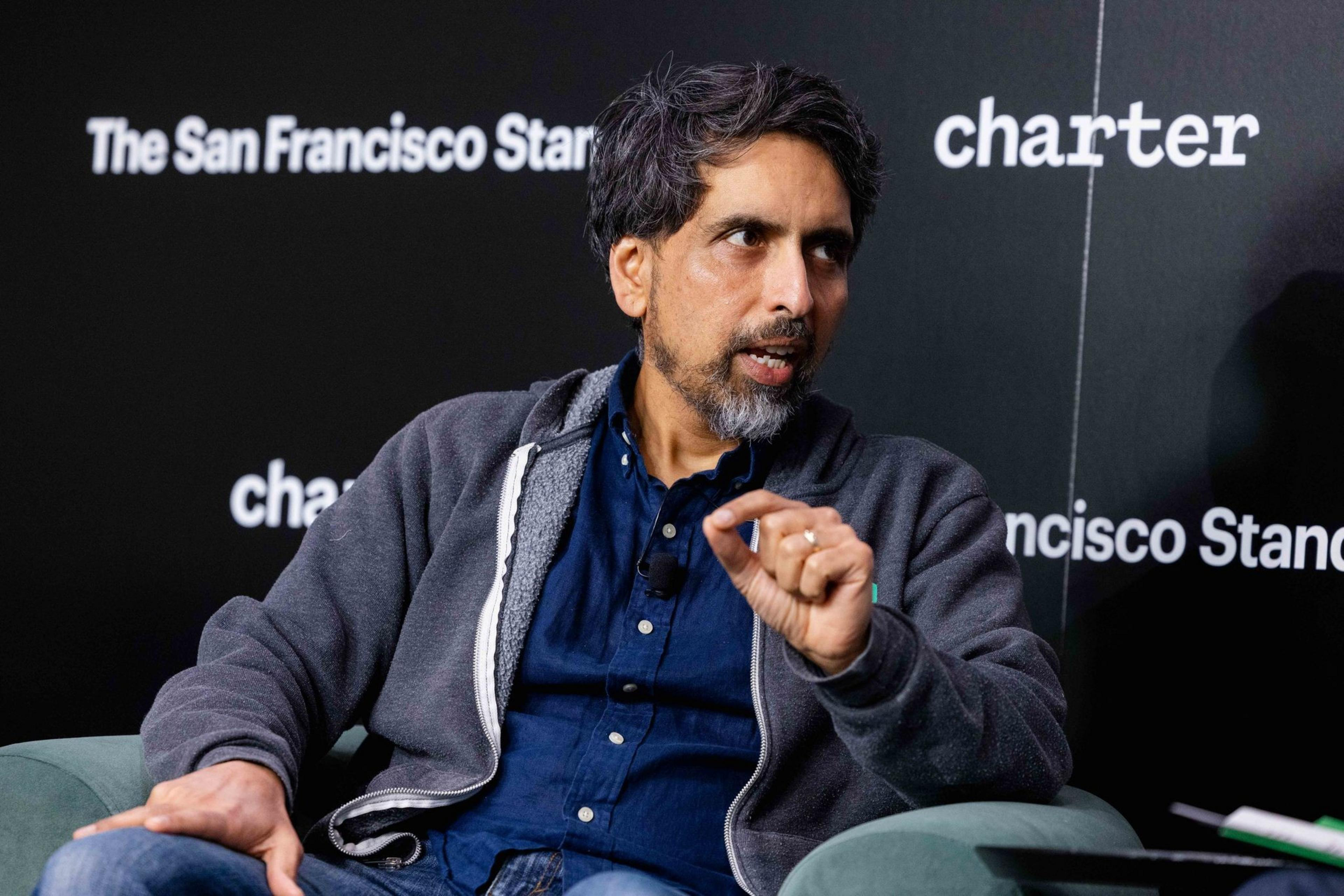 A man with graying hair and beard wears a blue shirt and gray hoodie, speaking with a hand gesture while seated against a black backdrop with text.
