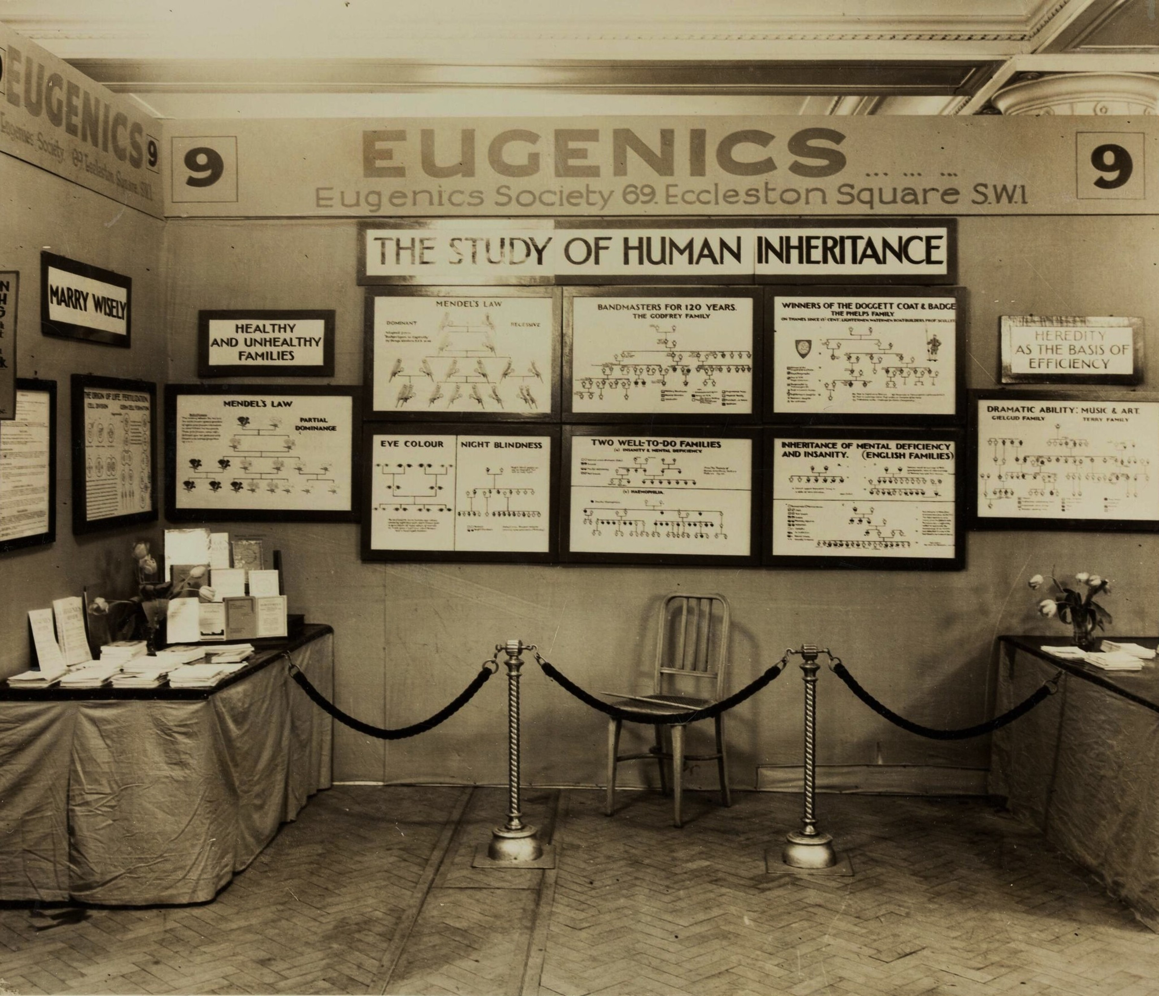 Posters and charts on human inheritance and eugenics are displayed on the walls of an exhibit room with tables and a wooden chair behind a rope barrier.