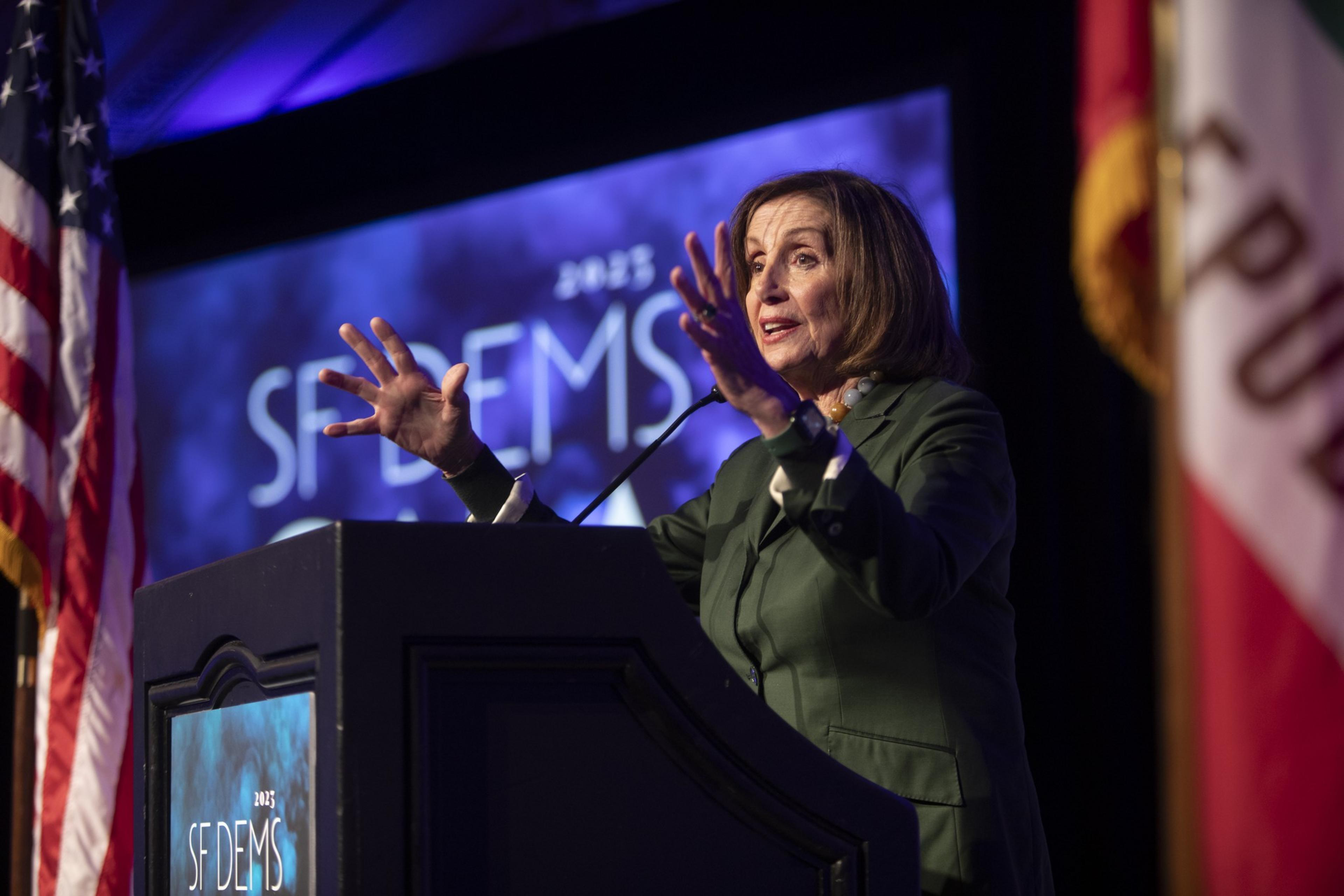 A woman is speaking at a podium with microphones, with the US flag to her left and event signage behind her.