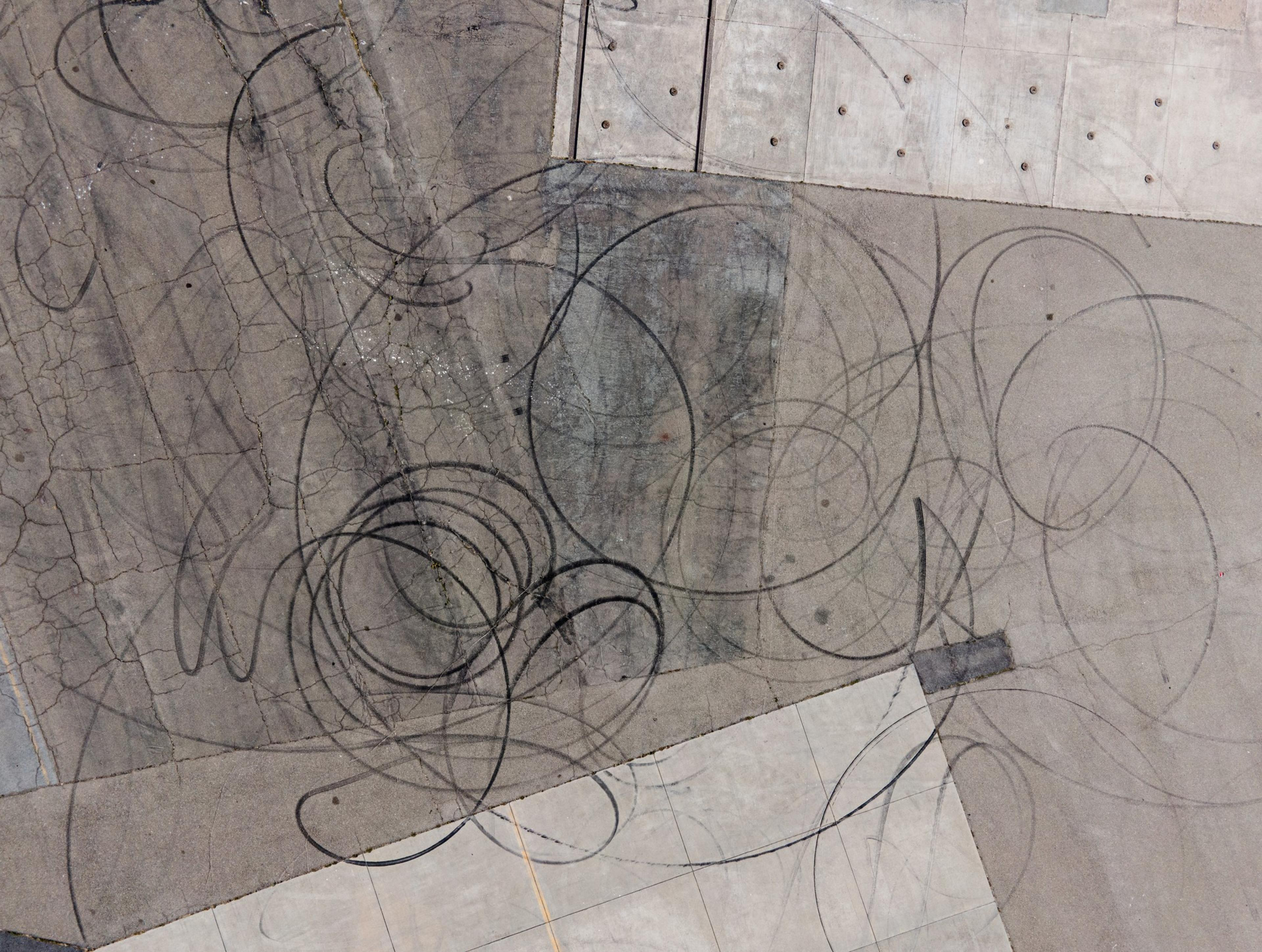 The image shows an aerial view of a concrete surface covered with circular tire skid marks, creating a chaotic pattern. The ground appears weathered and cracked.