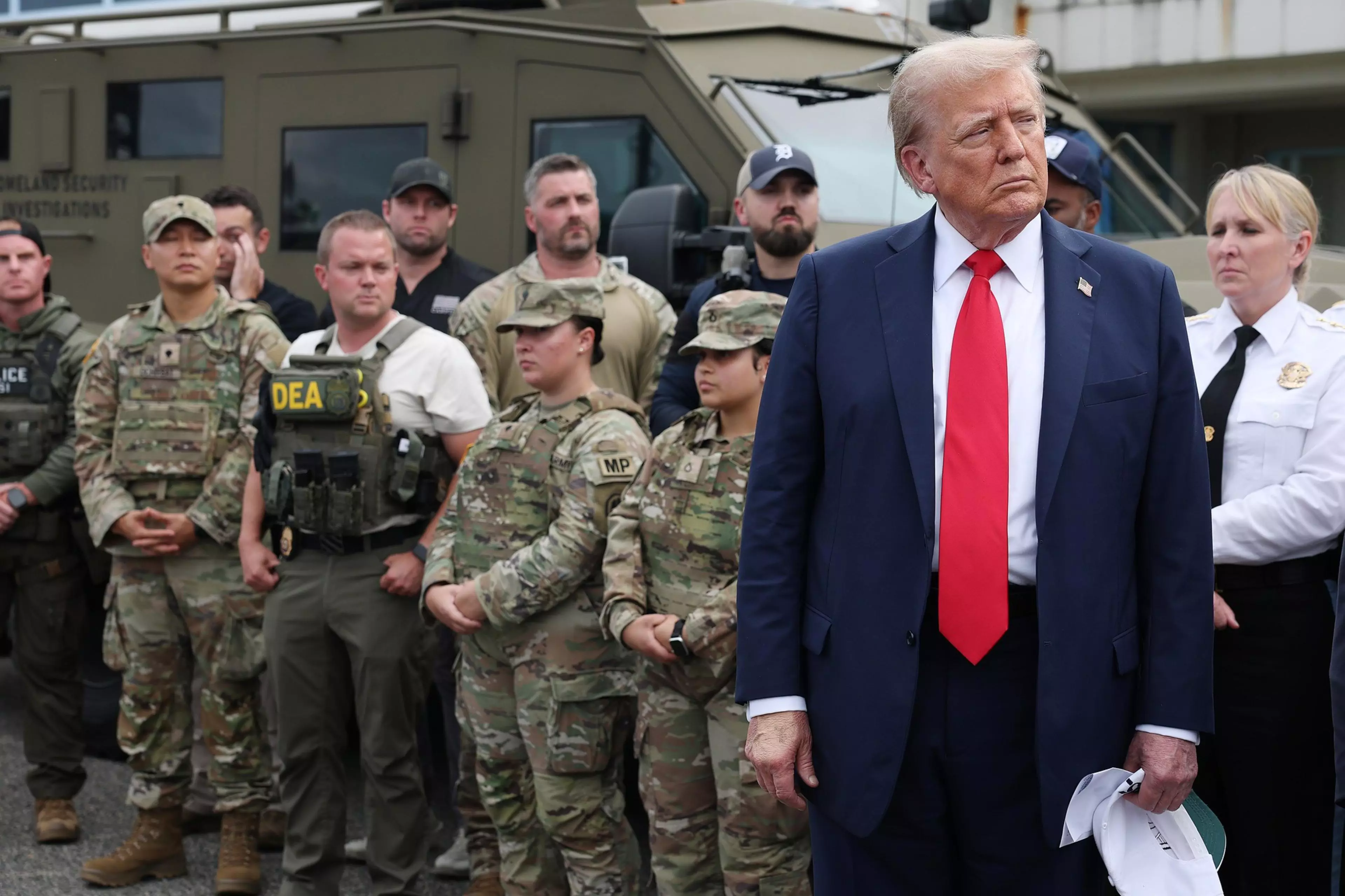 A man in a navy suit and red tie stands apart while various uniformed military and law enforcement personnel stand in a line behind him outdoors.