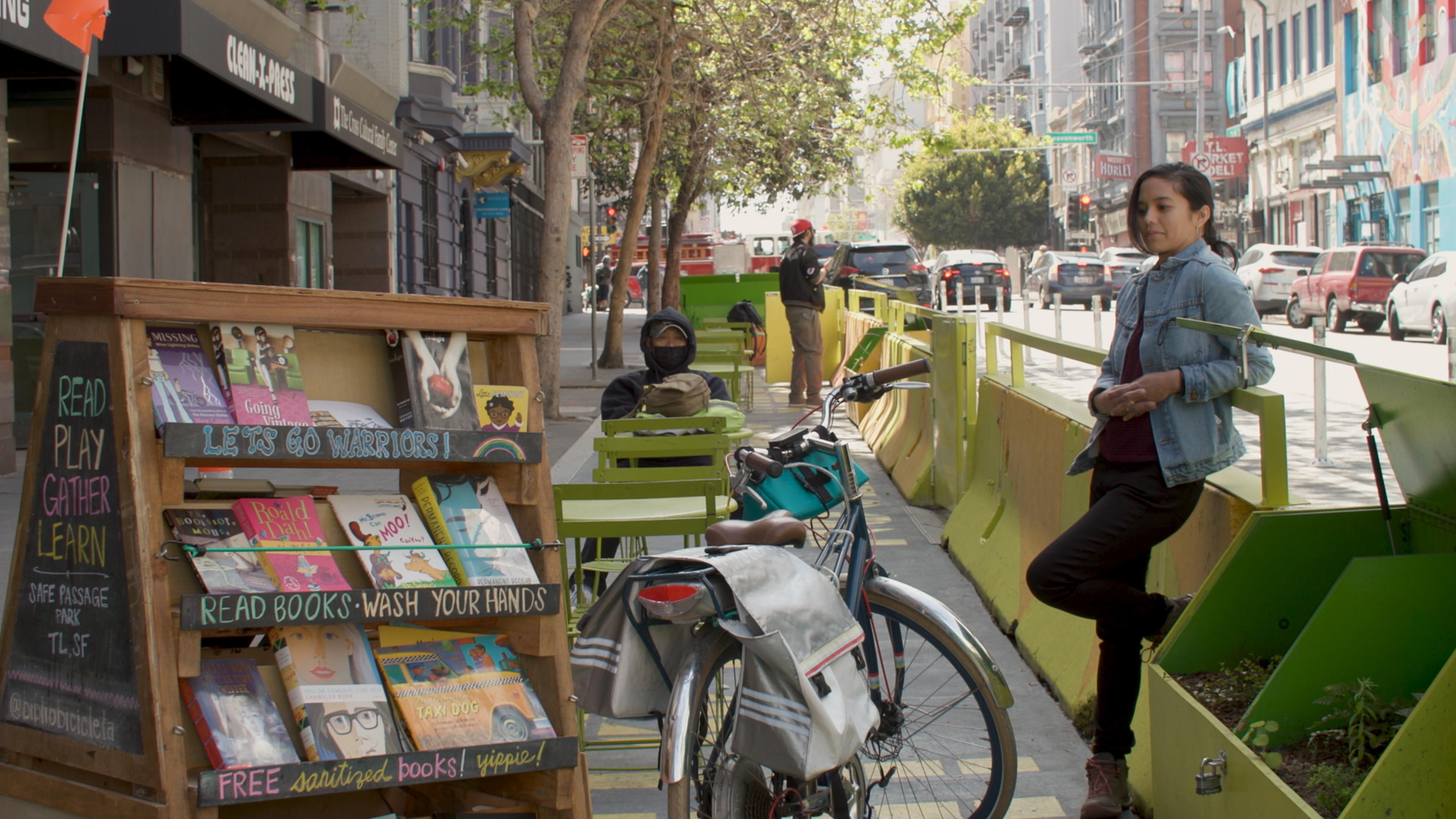 A street sidewalk features a small outdoor reading area with bookshelves, green tables and chairs, a bicycle, and a woman leaning against a green planter.