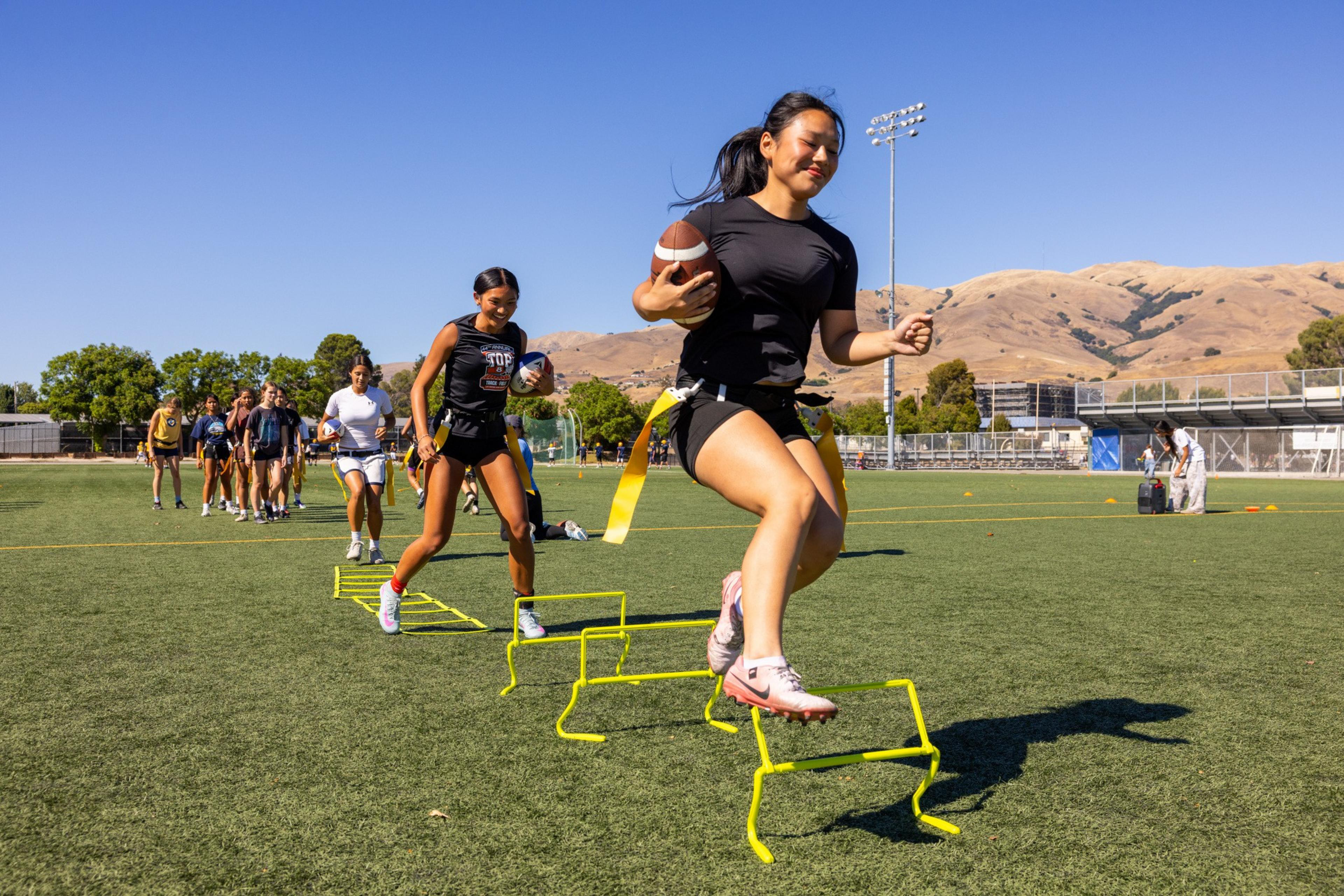 A group of young women run and jump over hurdles on a football field, with one holding a football and wearing a flag belt under clear skies.