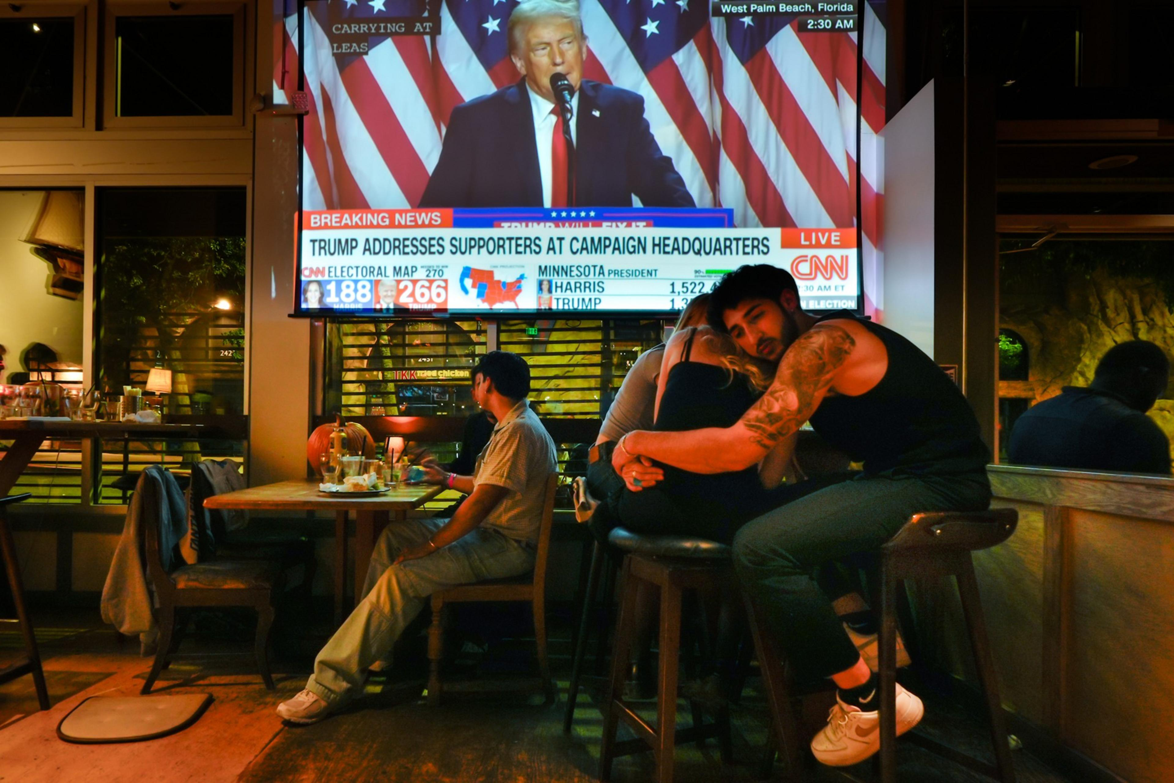 People are in a dimly lit bar watching a broadcast of a political speech. One person is seated at a table, while another person embraces someone at the bar.