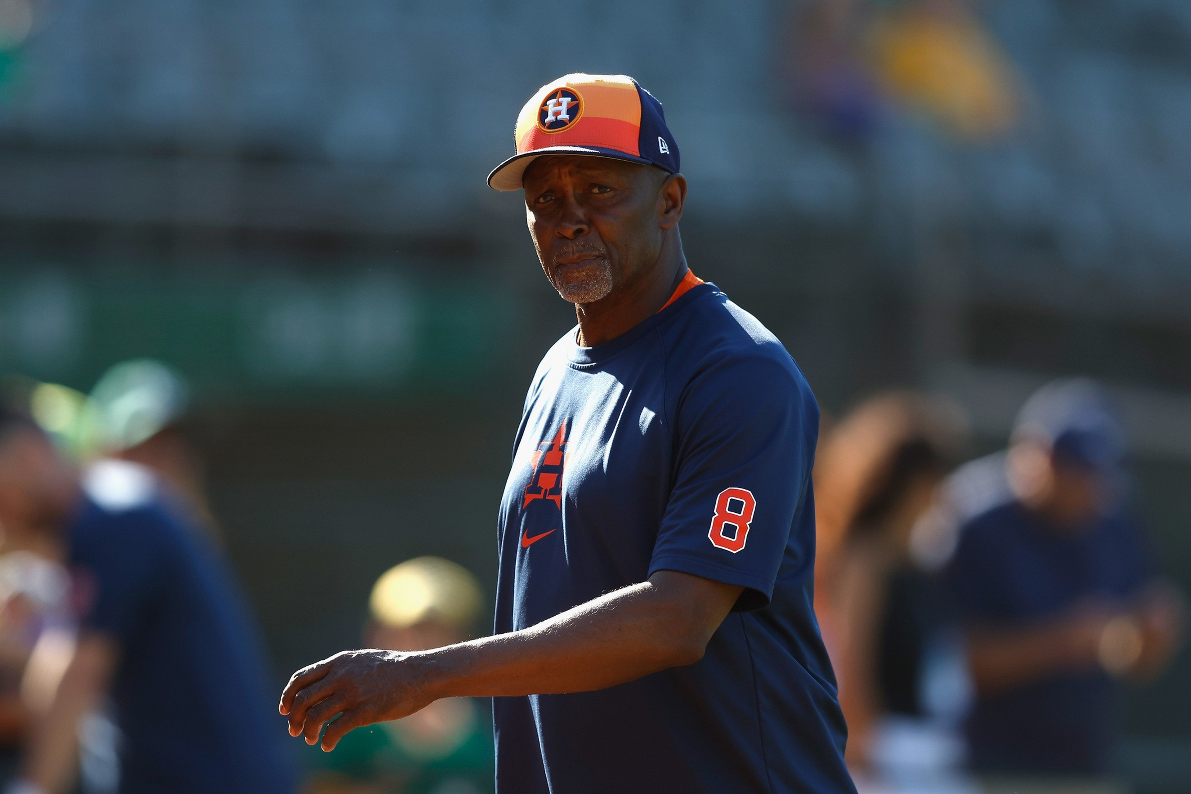 A man in a Houston Astros baseball shirt and cap stands outdoors, looking forward with a serious expression, surrounded by blurred people.