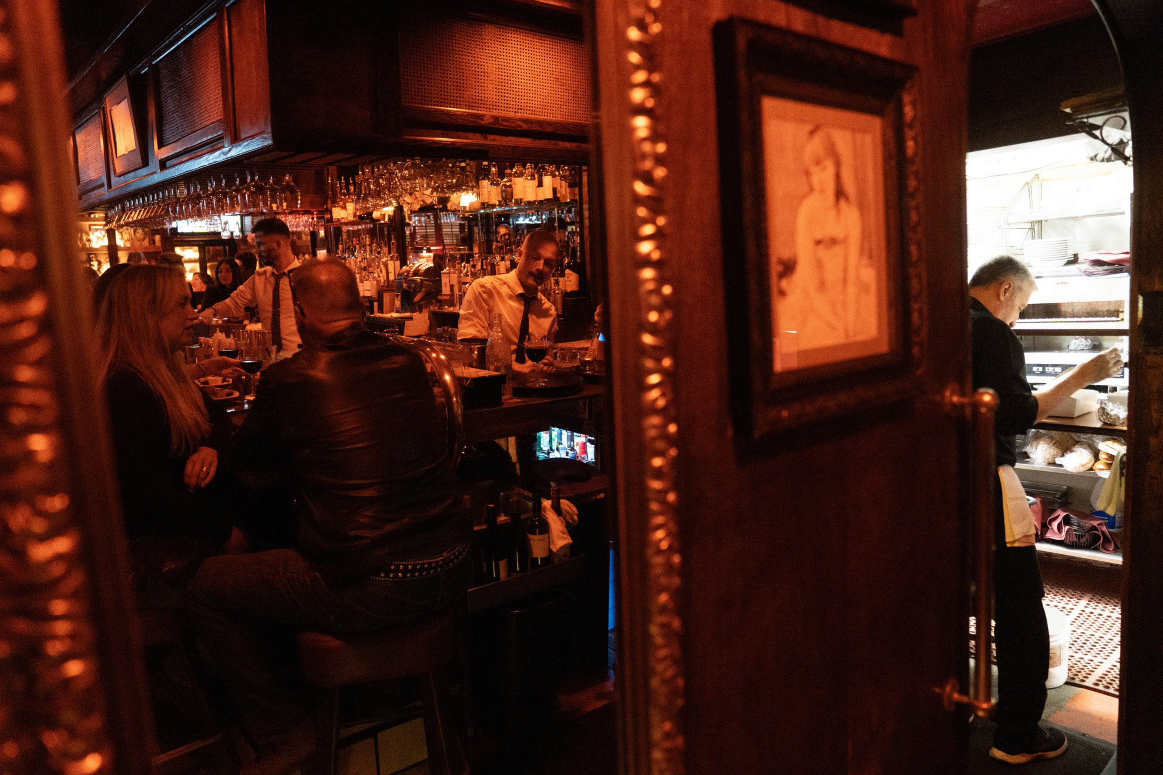 A dimly lit bar with people seated and a bartender behind the counter, next to a door showing a chef working in a kitchen.