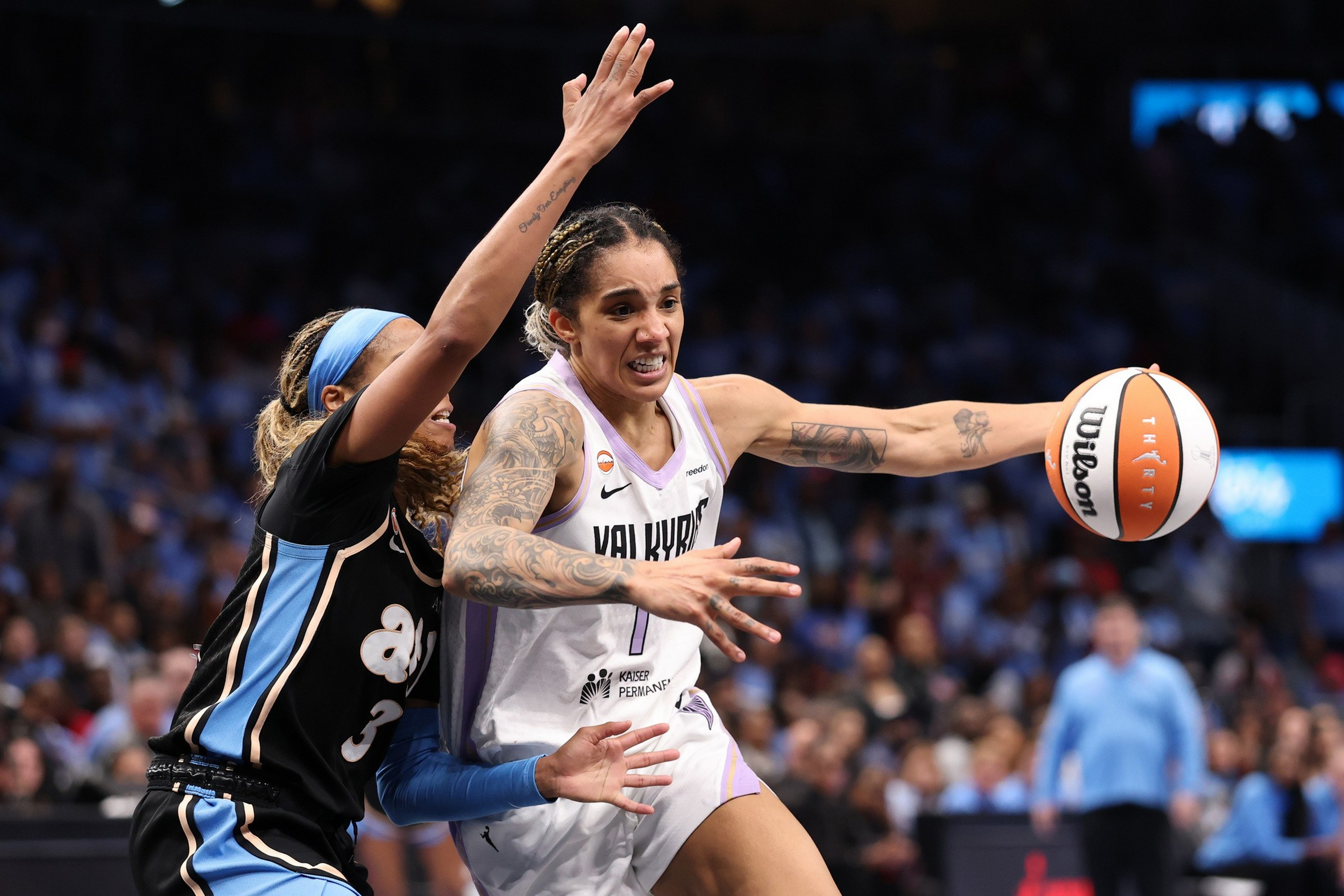 A basketball player in a white jersey aggressively drives past a defender in a black jersey, reaching out with the ball in her right hand.