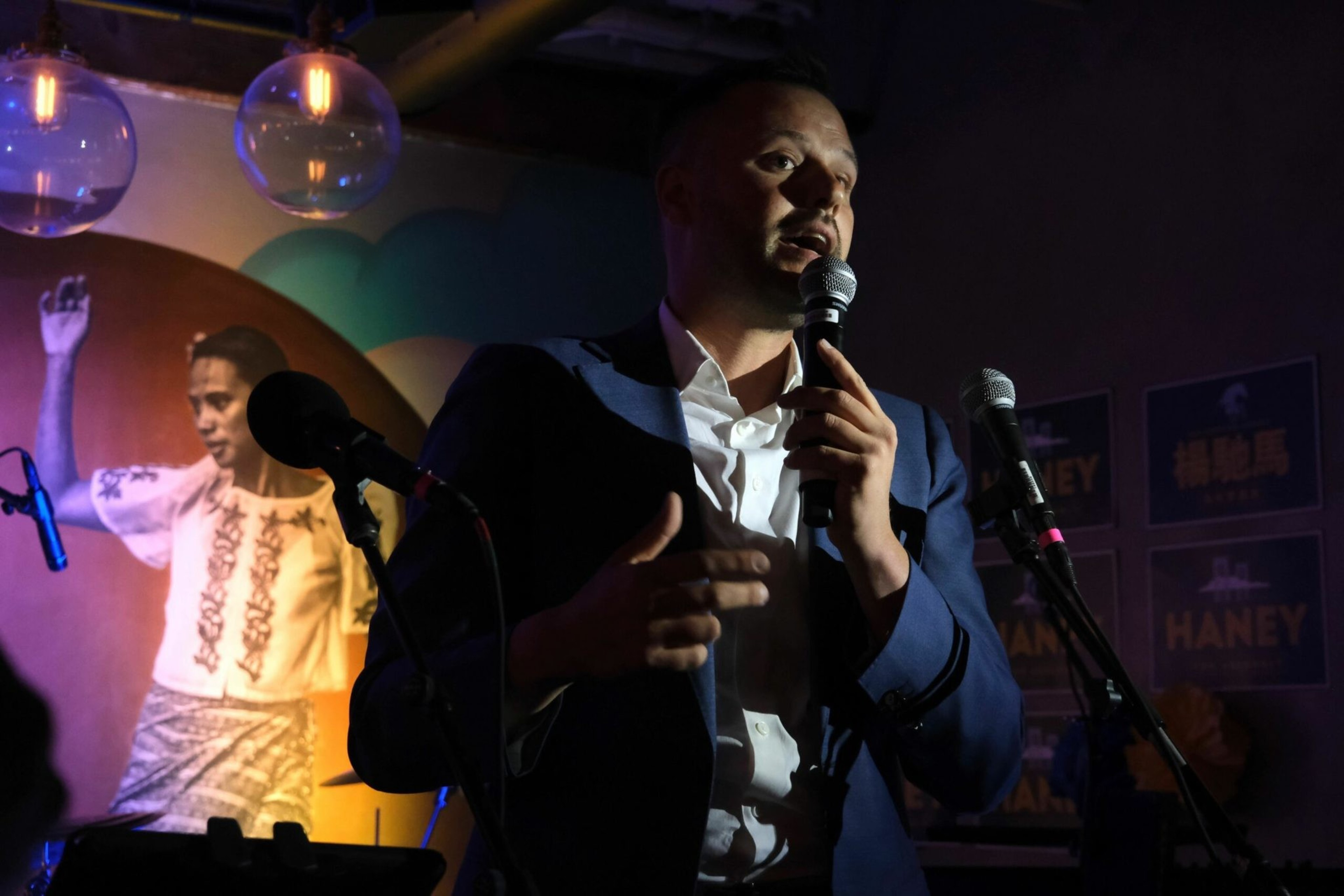 A man in a suit speaks into a microphone on a dimly lit stage with another microphone nearby and a colorful backdrop featuring a person in traditional attire.