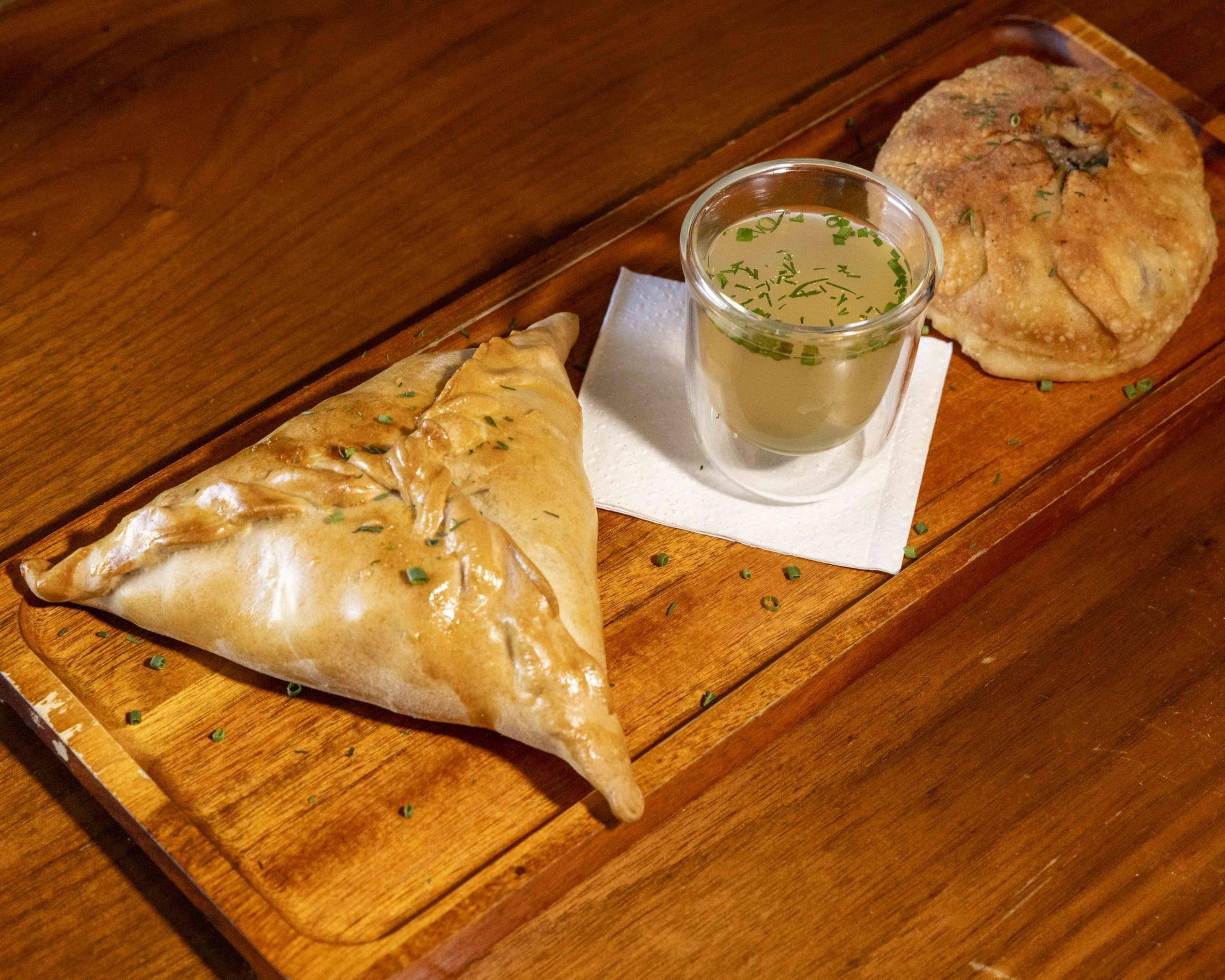 The image shows a wooden tray with a triangular pastry, a glass of clear soup garnished with herbs on a napkin, and a round, golden-brown flatbread.