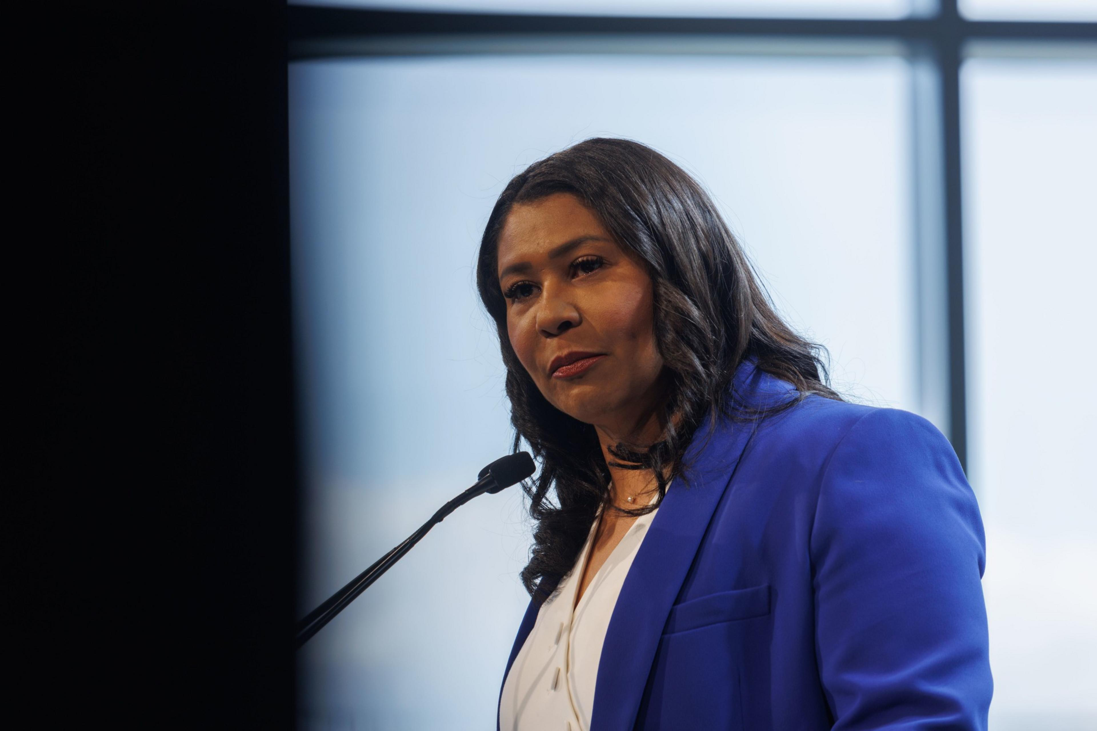 A woman in a blue blazer speaks at a podium, with large windows and soft light in the background.